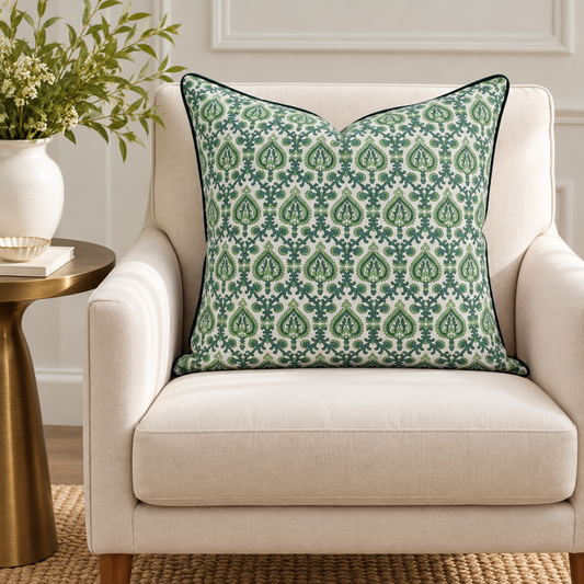 House of Mulmul - Blockprinted Cushion Covers-Pillow Cases. A square throw pillow with a green and white damask pattern and dark green piping, resting on a cream-colored armchair next to a gold side table with a white vase of greenery.