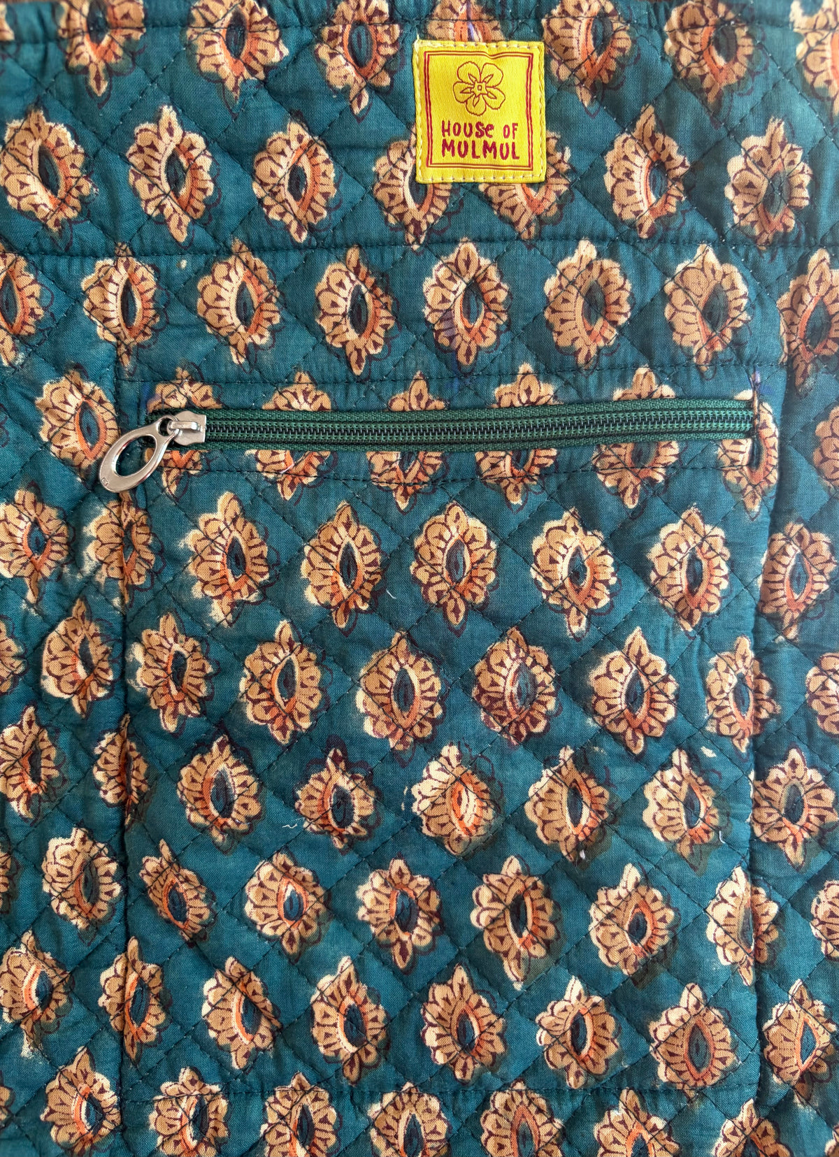 House of Mulmul - Bucket Tapered Tote Bag. Close-up of a quilted jacket or bag featuring a teal blue background with a repeating tan and brown floral block print pattern, a horizontal zipper pocket, and a yellow brand label reading 'House of Moolool'.