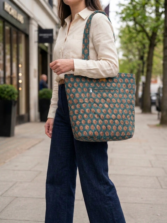 House of Mulmul - Bucket Tapered Tote Bag. A woman wearing a cream shirt and dark blue wide-leg jeans carries a large tote bag with a blue and beige floral pattern on a city sidewalk.