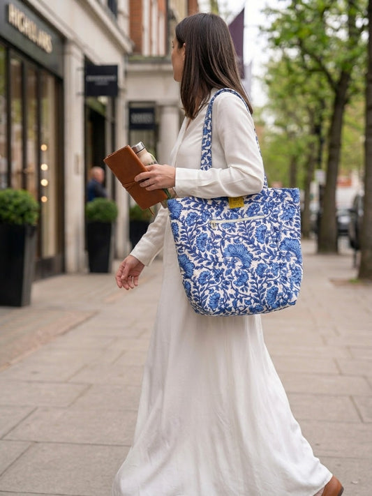 House of Mulmul - Bucket Tapered Tote Bag. A woman in a white dress walks on a city sidewalk carrying a large blue and white floral print tote bag over her shoulder.