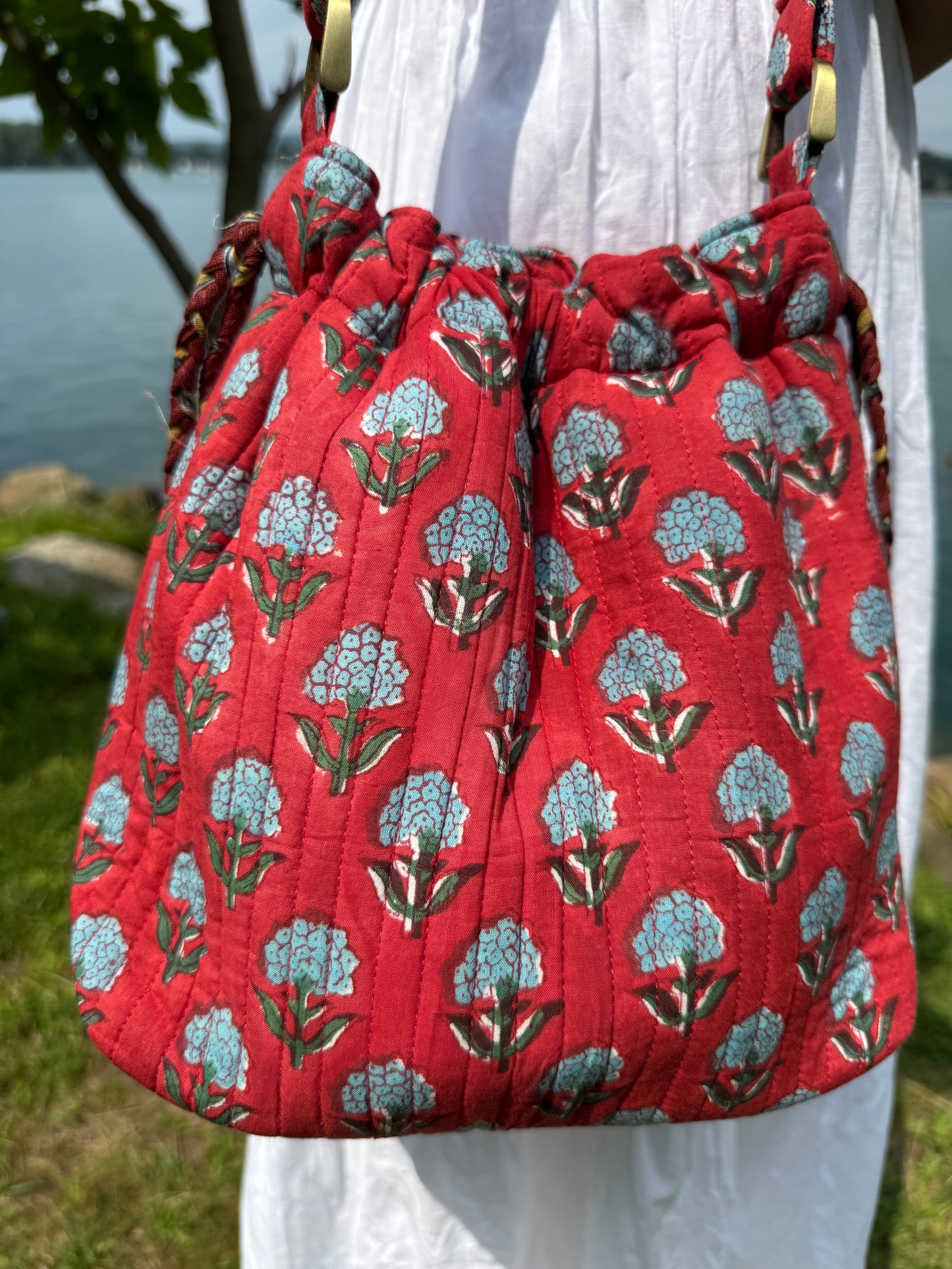 House of Mulmul - Garden Wayfarer Bucket Bag, Cotton bag, Crossbody sling bag. Red drawstring bag with floral pattern held by a person outdoors.