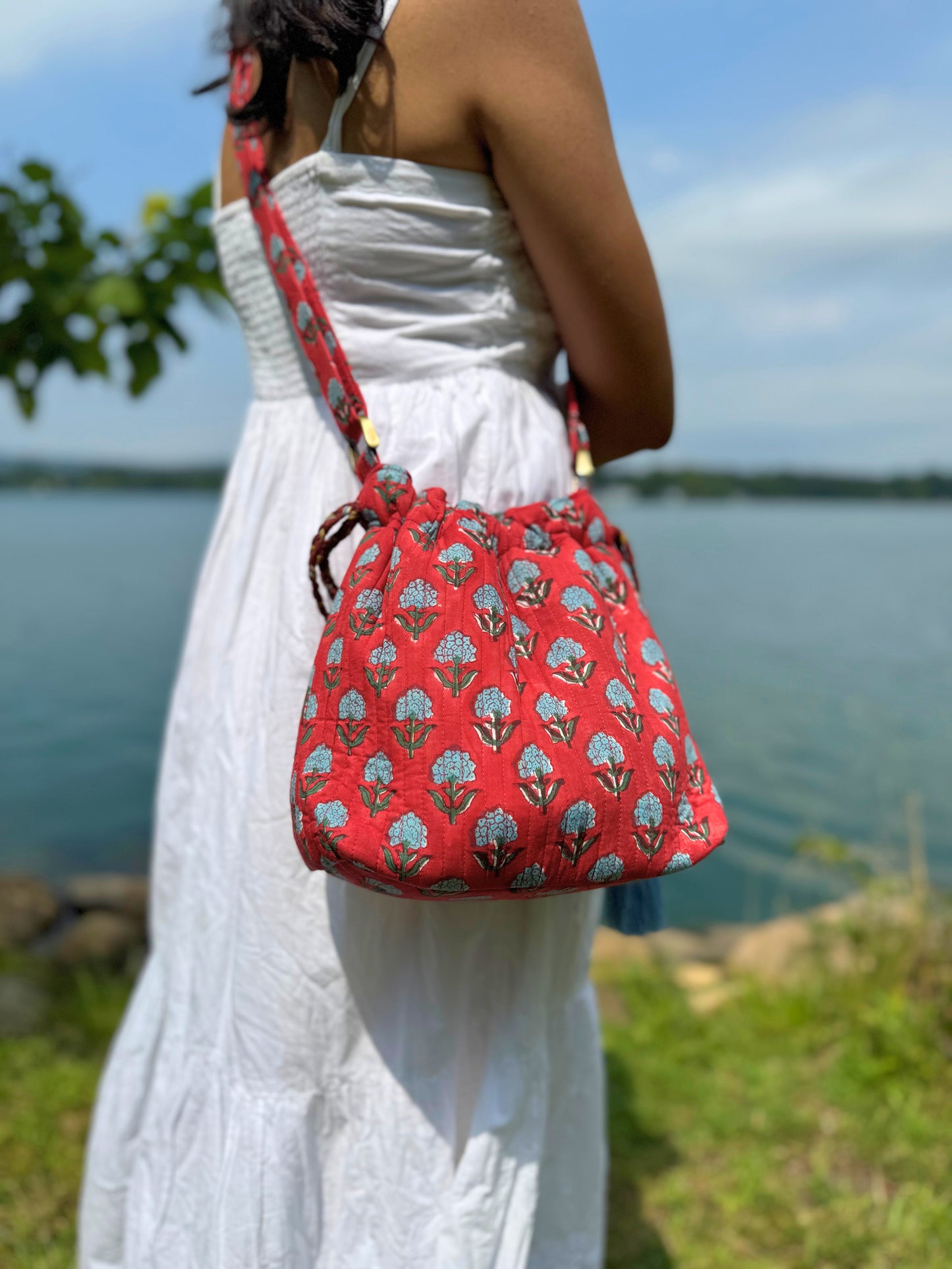 House of Mulmul - Garden Wayfarer Bucket Bag, Cotton bag, Crossbody sling bag. Person wearing a white dress with a red patterned bag by a body of water
