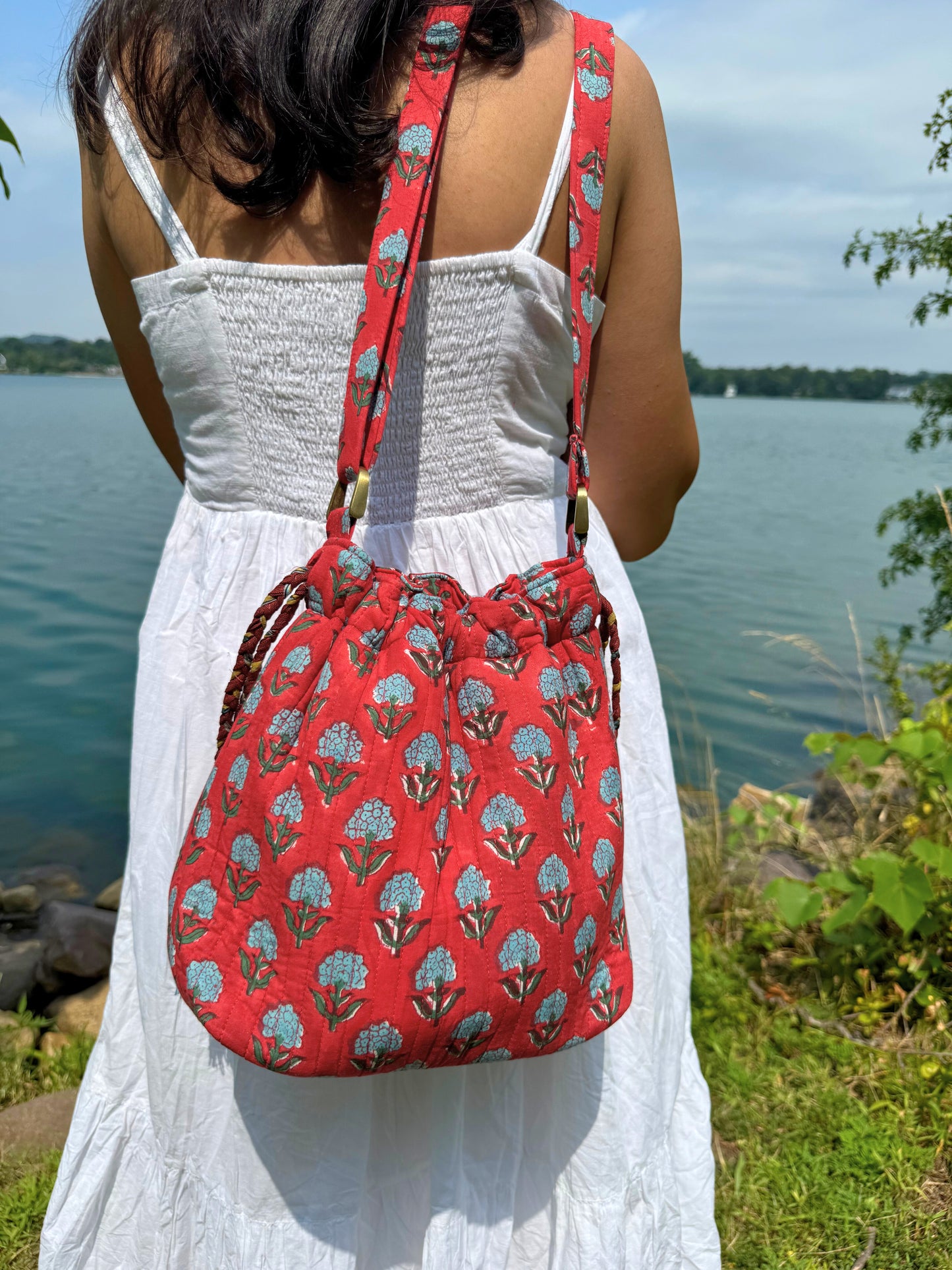 House of Mulmul - Garden Wayfarer Bucket Bag, Cotton bag, Crossbody sling bag. Person wearing a red floral-patterned bag by a body of water