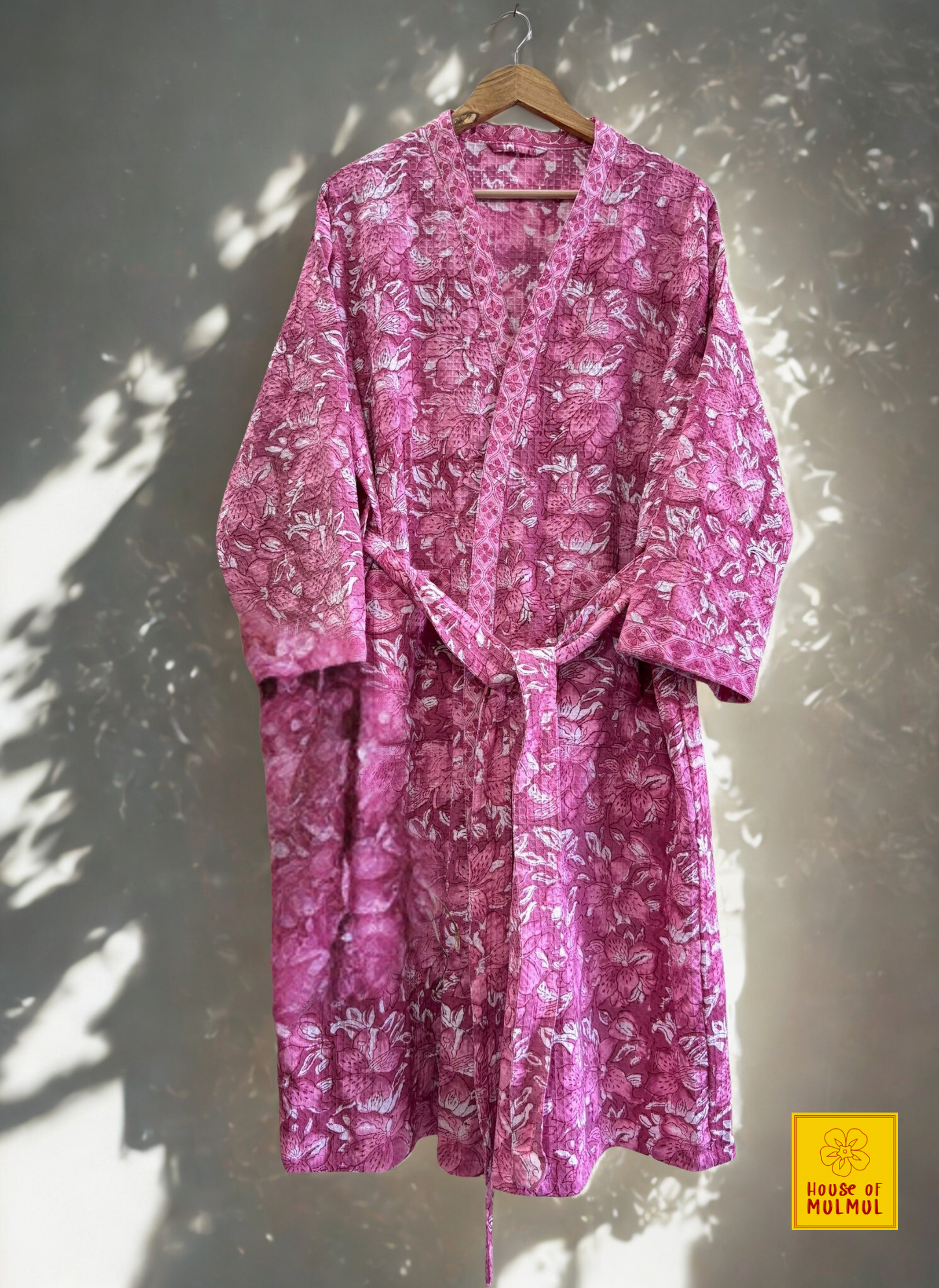 House of Mulmul - Blockprinted Cotton Bathrobe. Pink floral robe hanging on a wooden hanger against a white wall.
