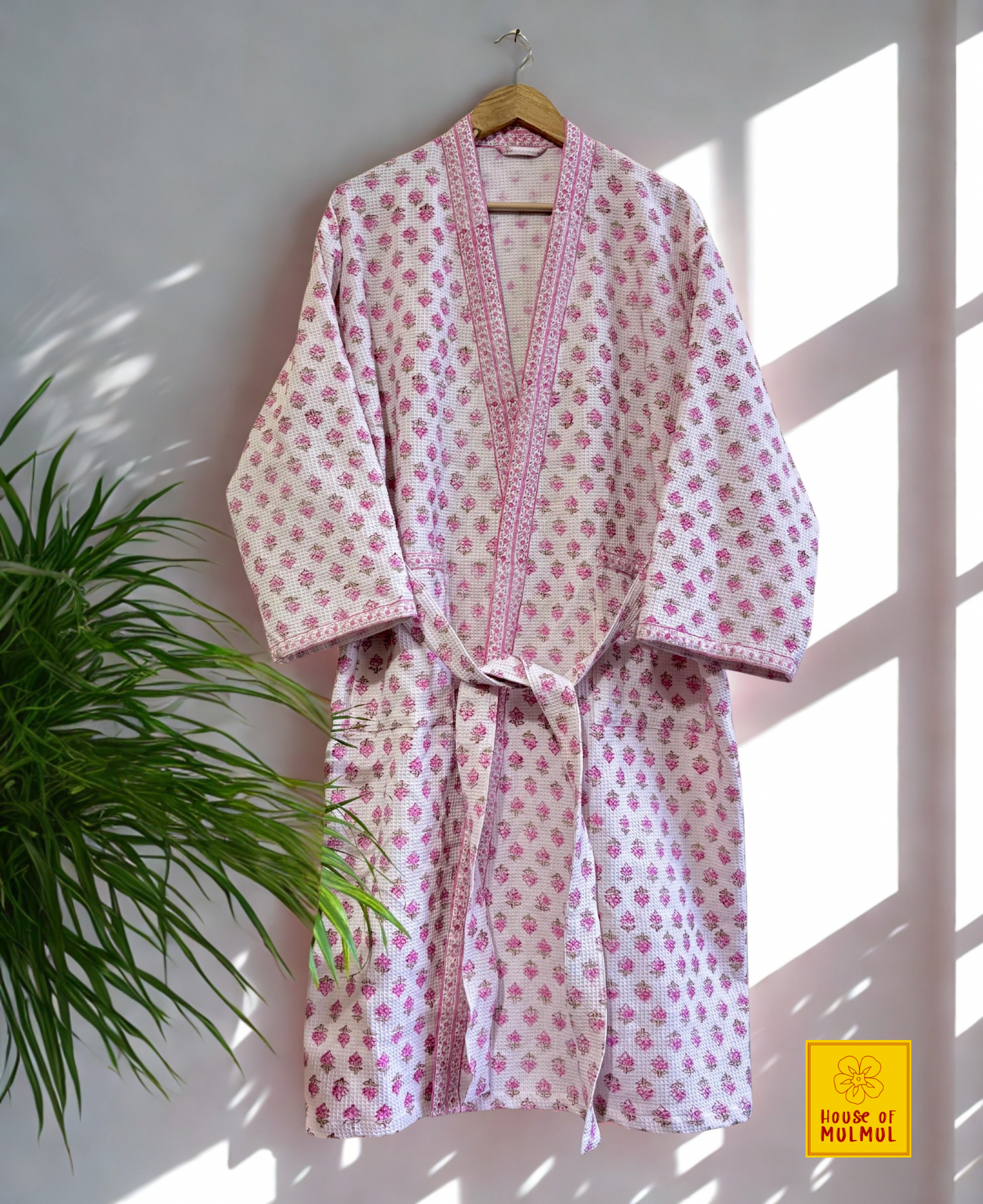 House of Mulmul - Blockprinted Cotton Bathrobe. Pink and white floral patterned robe hanging on a wooden hanger against a white wall with natural light.