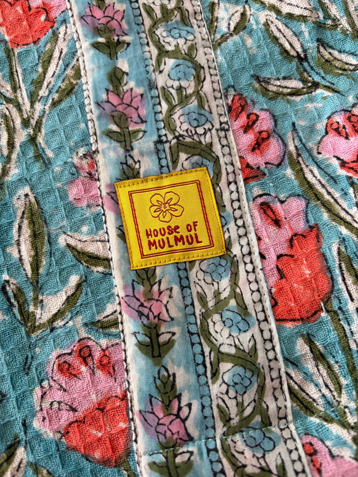 House of Mulmul - Blockprinted Cotton Bathrobe. A close-up of a colorful floral bathrobe with vertical patterned bands and a yellow square patch reading 'HOUSE OF MULMUL'.