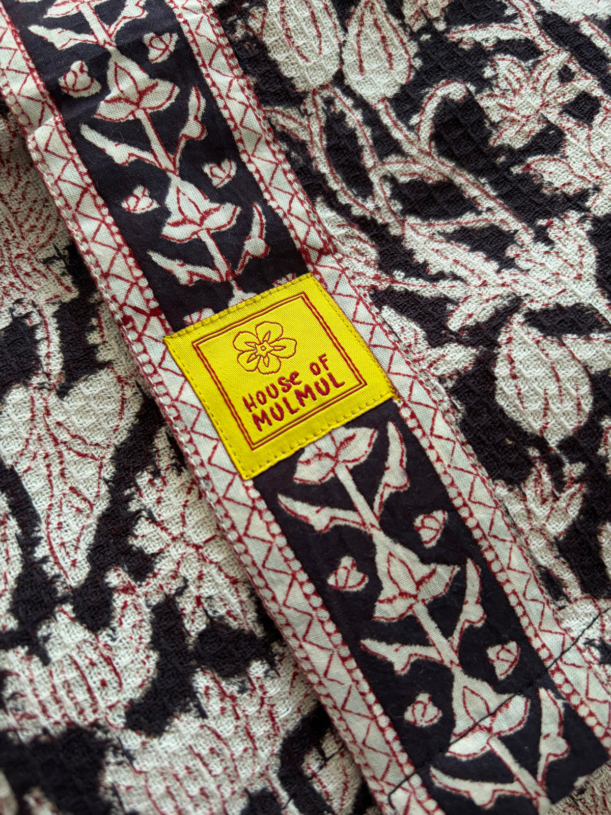 House of Mulmul - Blockprinted Cotton Bathrobe. A close-up of a patterned bathrobe with a bright yellow woven label reading 'HOUSE OF MUMUL'. The fabric has a dark black and cream floral block-print style pattern with red stitched borders.
