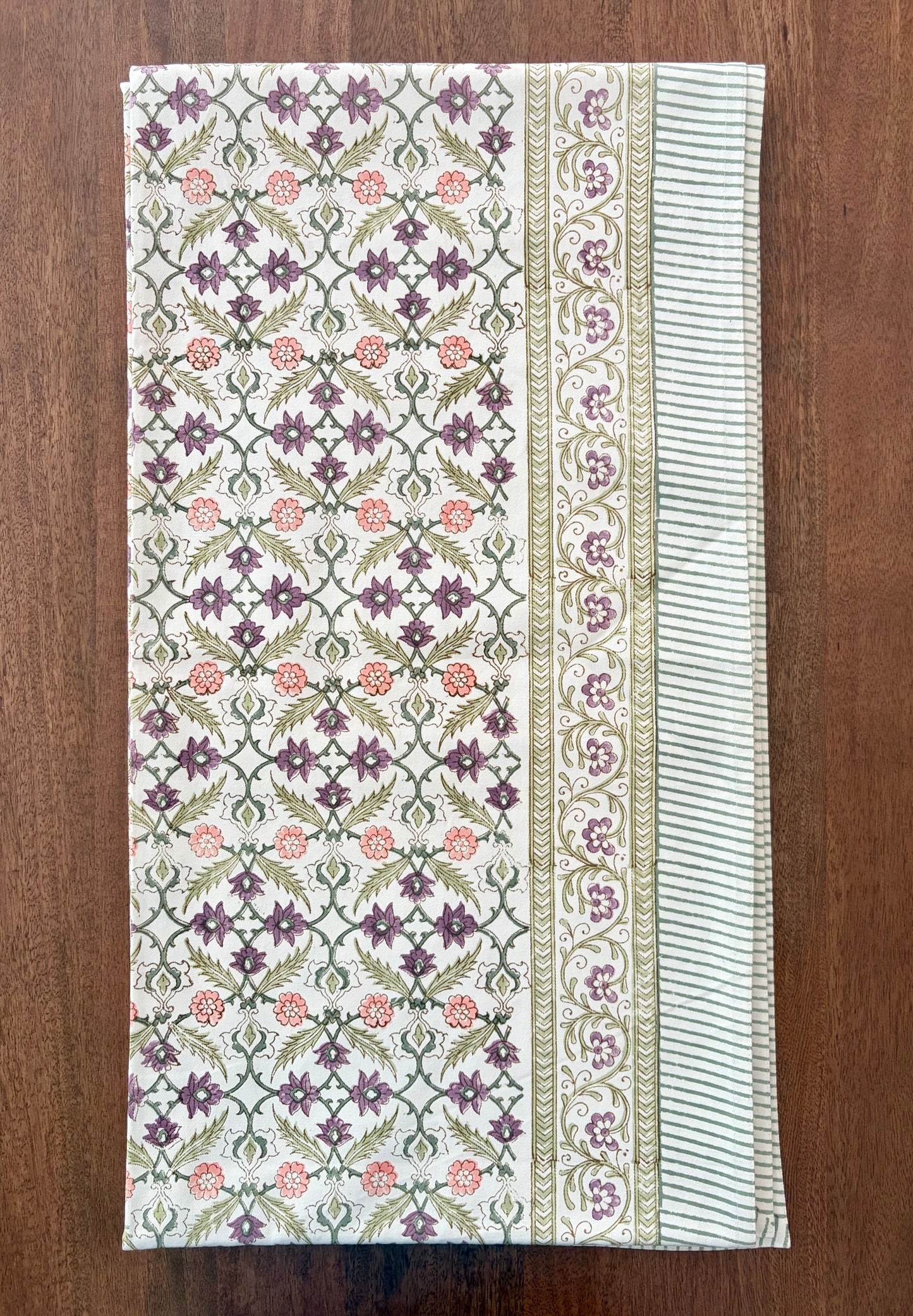 House of Mulmul - Blockprinted Cotton Table Cloth. A rectangular cotton table runner with an intricate block-printed floral pattern, laid flat on a dark brown wooden surface. The runner features a central panel with purple and pink flowers on a green and white geometric lattice, flanked by two borders with striped and floral details.