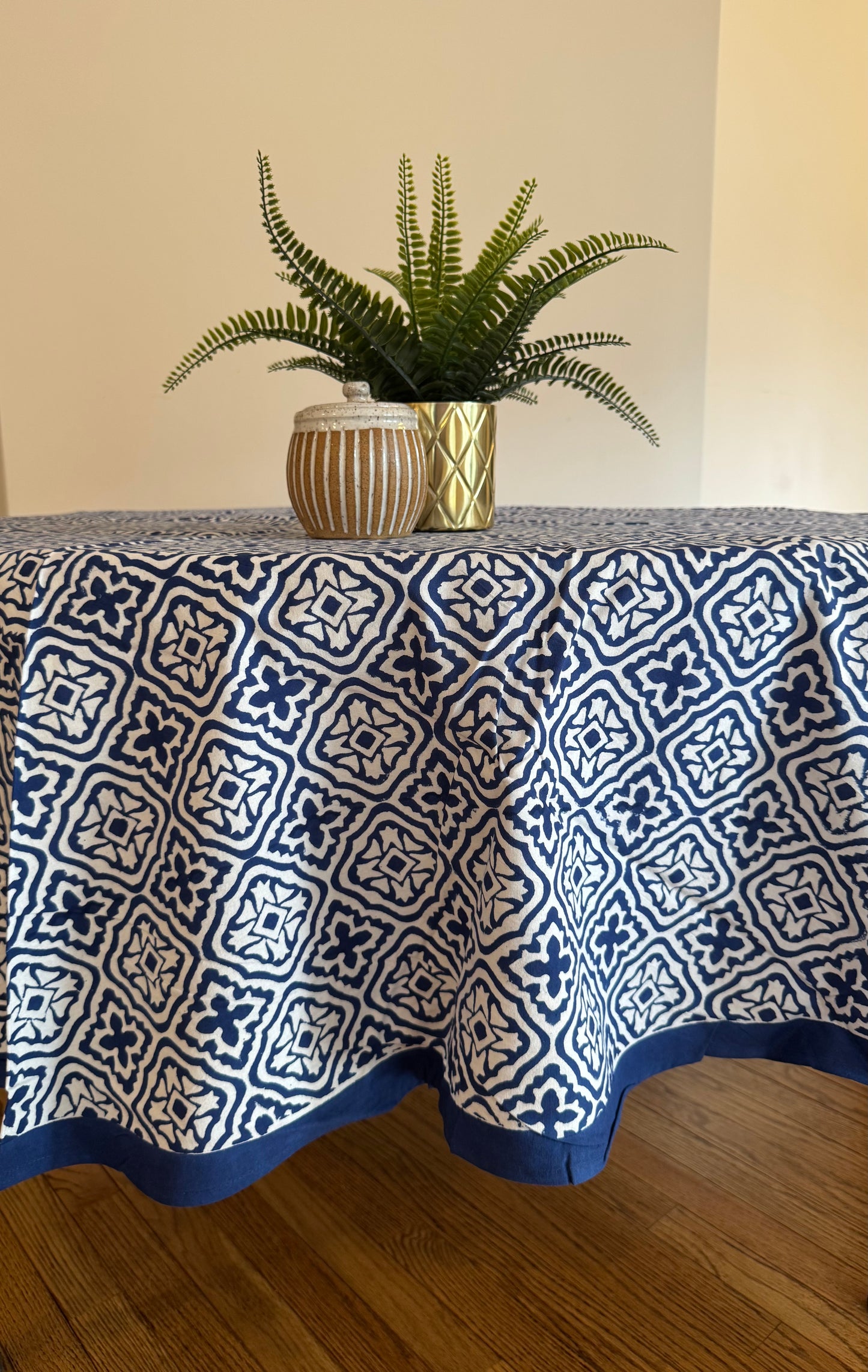 House of Mulmul - Cotton Table Cloth Round. Tablecloth with blue and white pattern on a table, with a plant in the background.