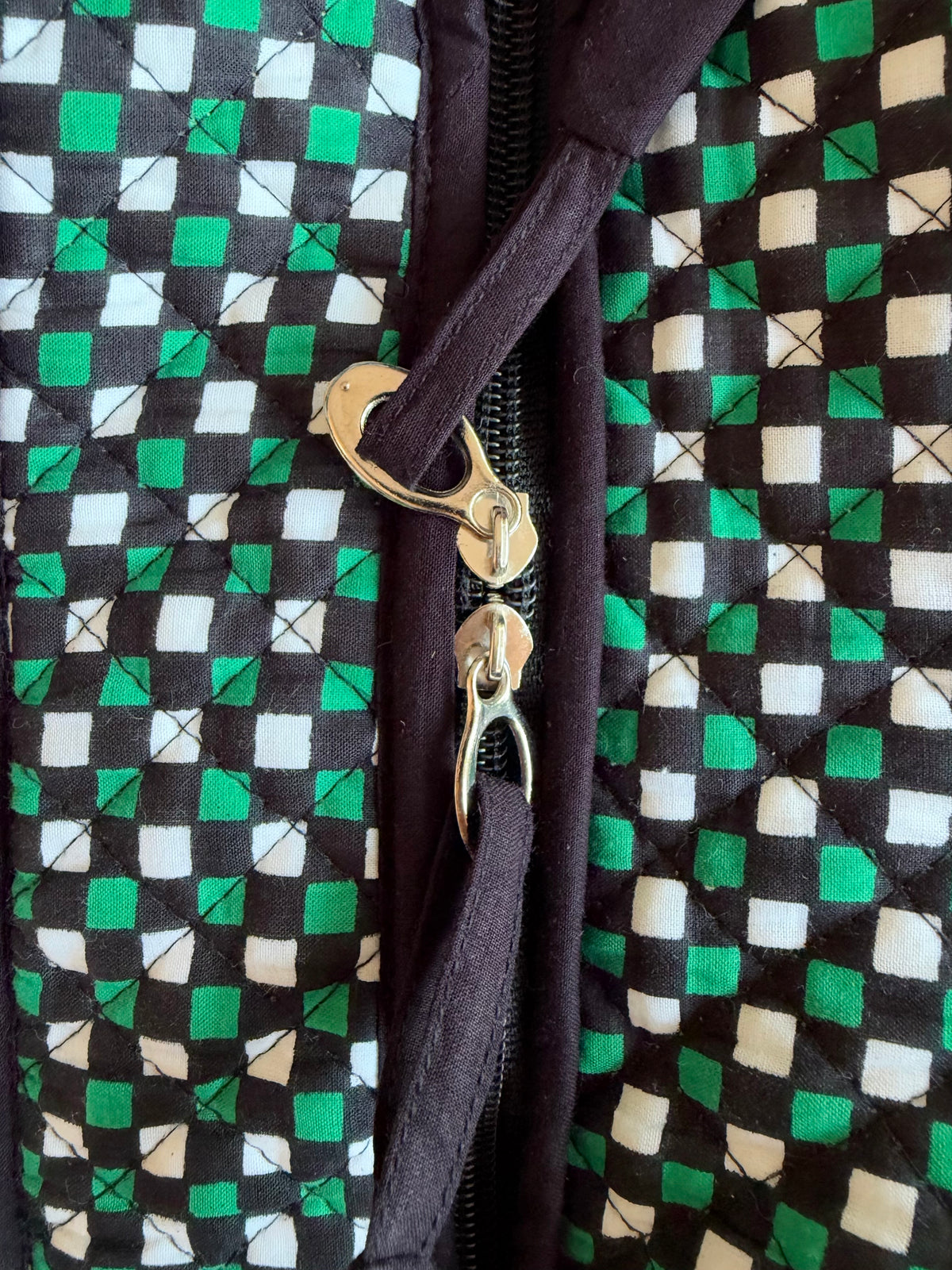 House of Mulmul - Cotton Tote Bags. Close-up of a quilted zippered jacket or bag with a black, green, and white checkered pattern on the exterior and a dark brown interior lining, featuring gold-tone metal zipper pulls.