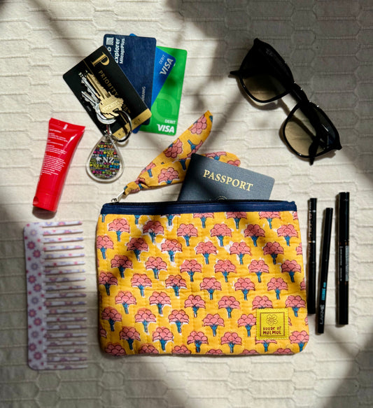 House of Mulmul - Quilted Flat Passport Pouch. Yellow patterned pouch with travel items including a passport, sunglasses, and toiletries on a textured surface.