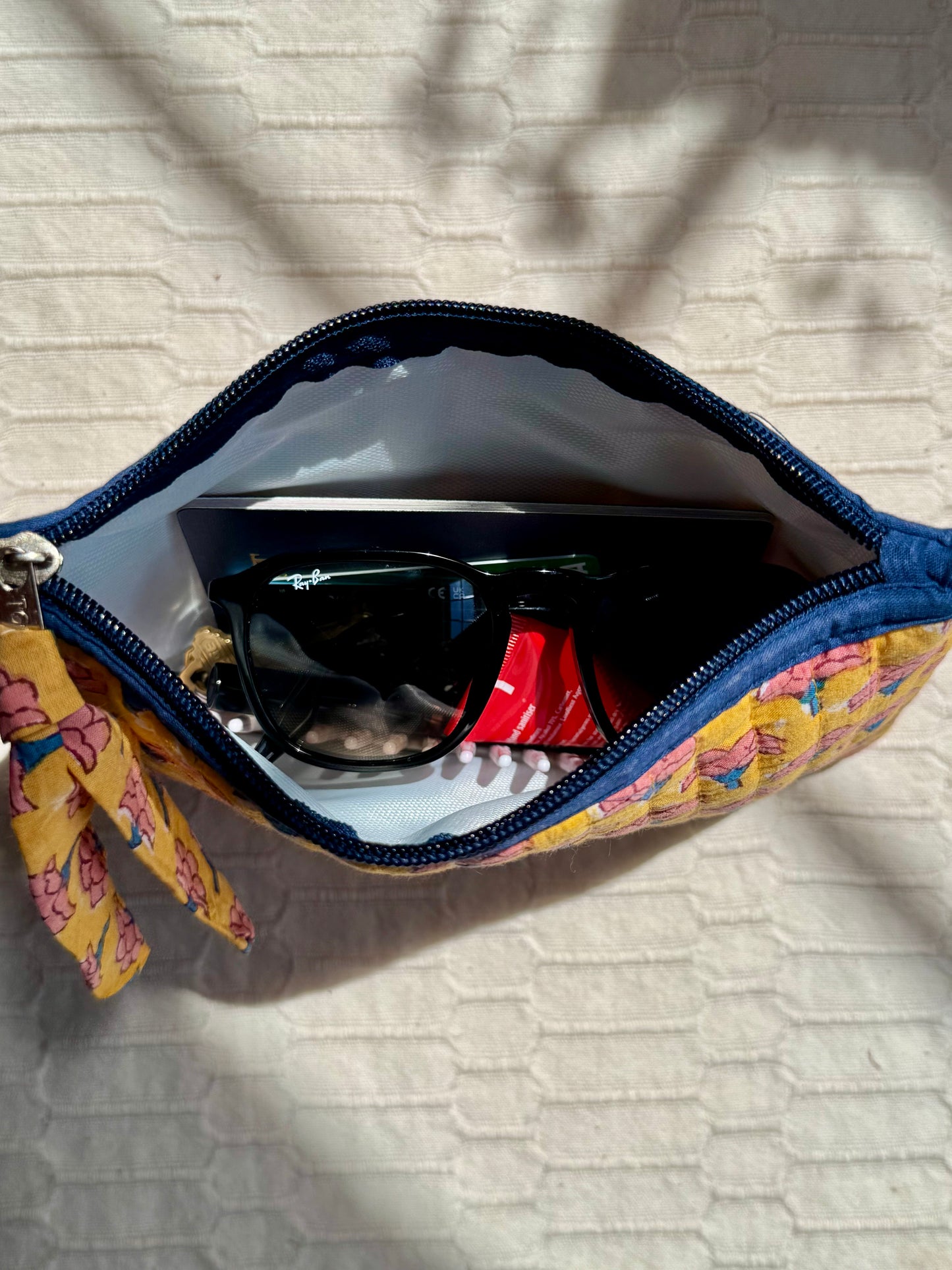House of Mulmul - Quilted Flat Passport Pouch. Floral-patterned pouch with sunglasses and a red item on a textured surface.