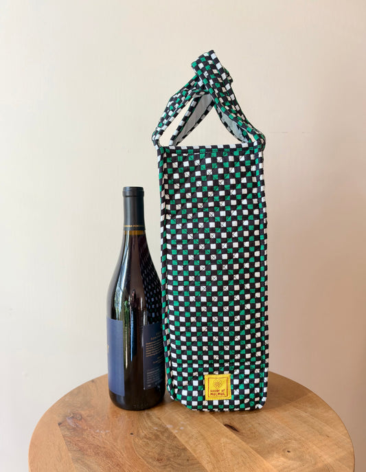 House of Mulmul - Quilted Wine Tote Bag. A tall fabric wine bottle carrier with a green, white, and black checkered pattern and a knotted top handle, shown next to a wine bottle on a round wooden stool against a plain beige wall.
