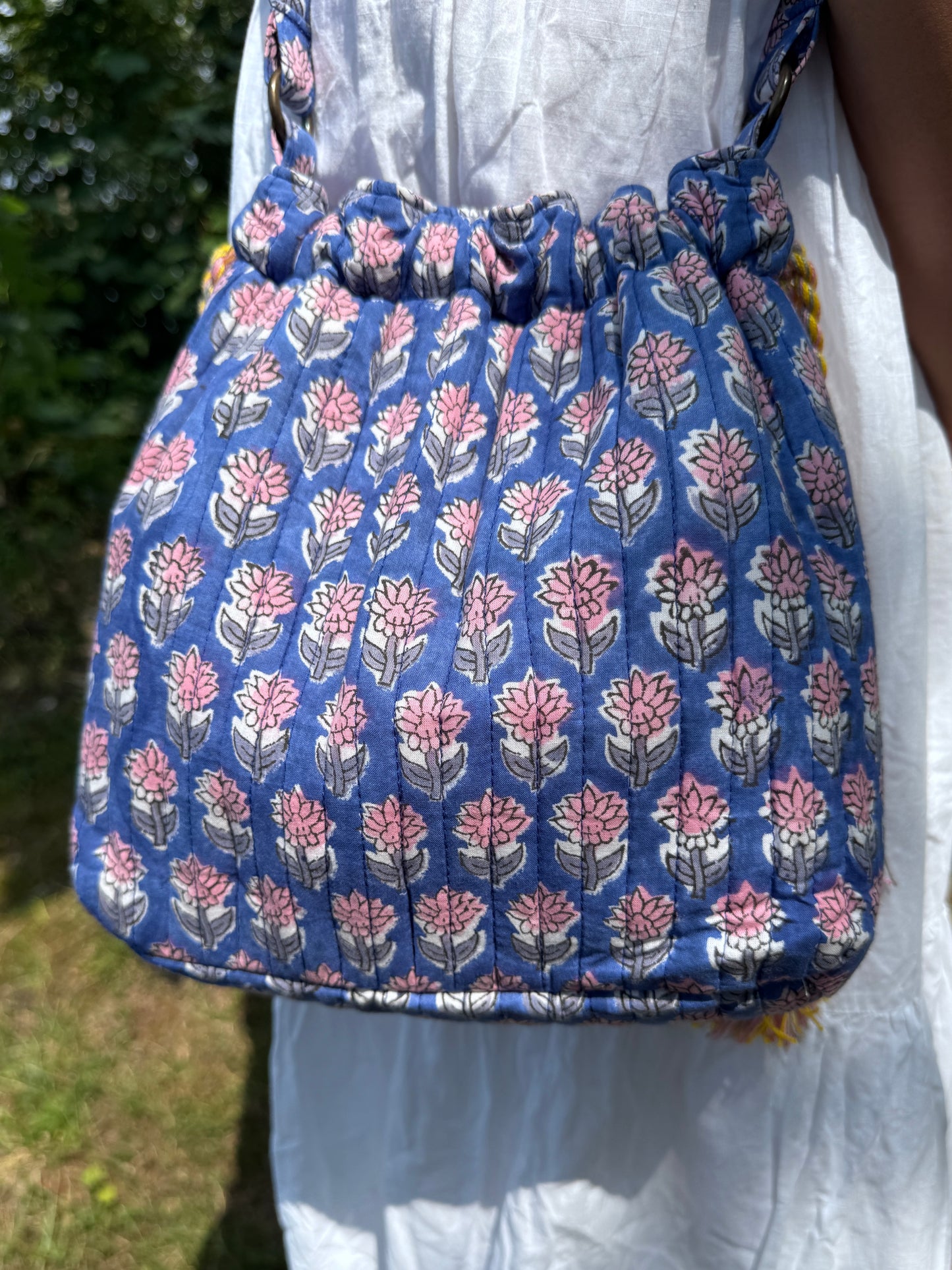 House of Mulmul - Garden Wayfarer Bucket Bag, Cotton bag, Crossbody sling bag. Blue floral-patterned bag held by a person wearing a white dress outdoors.