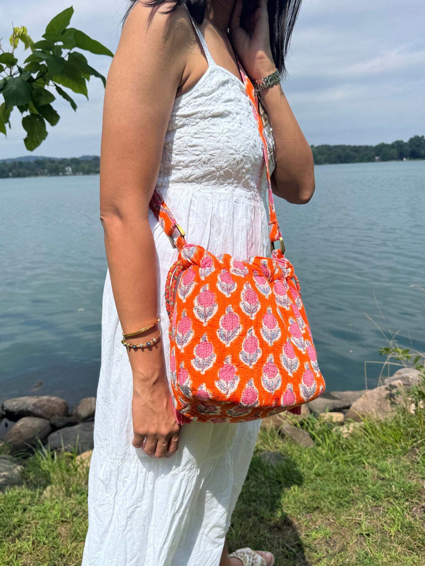 House of Mulmul - Garden Wayfarer Bucket Bag, Cotton bag, Crossbody sling bag. Person wearing a white dress with an orange patterned bag by a body of water.