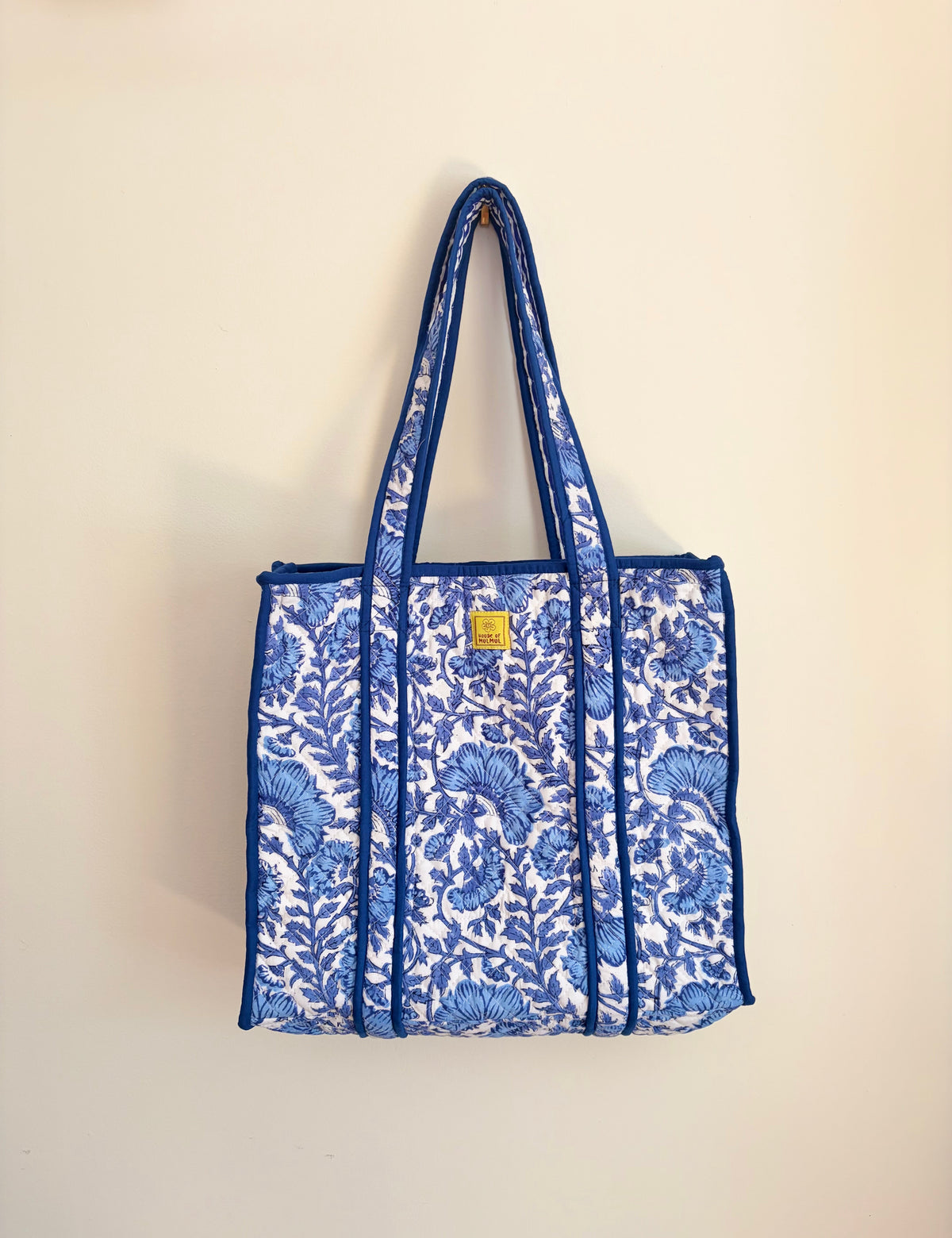 House of Mulmul - Blockprinted Cotton Tote Bag, Travel Bag. A blue and white floral print tote bag with blue handles and trim, hanging against a plain beige wall.