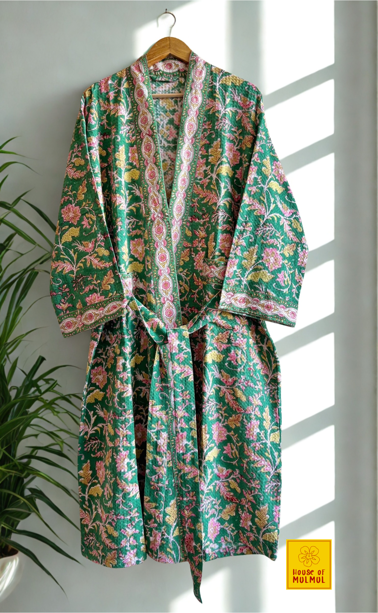 House of Mulmul - Blockprinted Cotton Bathrobe. Floral patterned cotton bath robe hanging on a wooden hanger against a white wall with a plant to the left.
