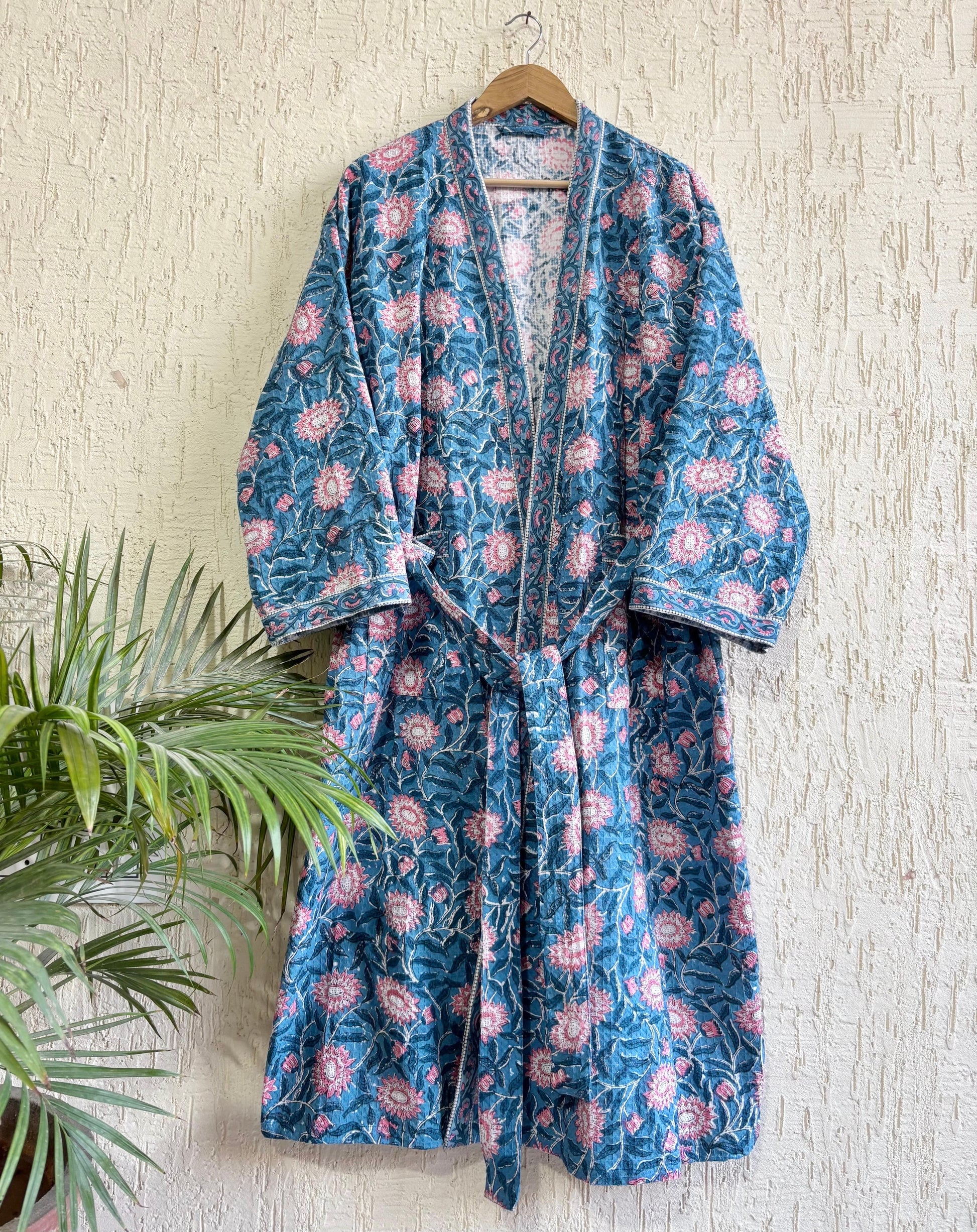 House of Mulmul - Blockprinted Cotton Bathrobe. Blue and pink floral patterned robe hanging on a wooden hanger against a textured wall.