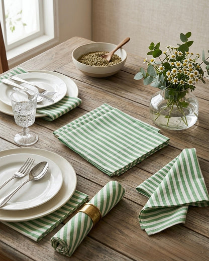 House of Mulmul - Blockprinted Cotton Table Napkins. Dining table set with green & white striped napkins, white plates, and silverware on a wooden surface.