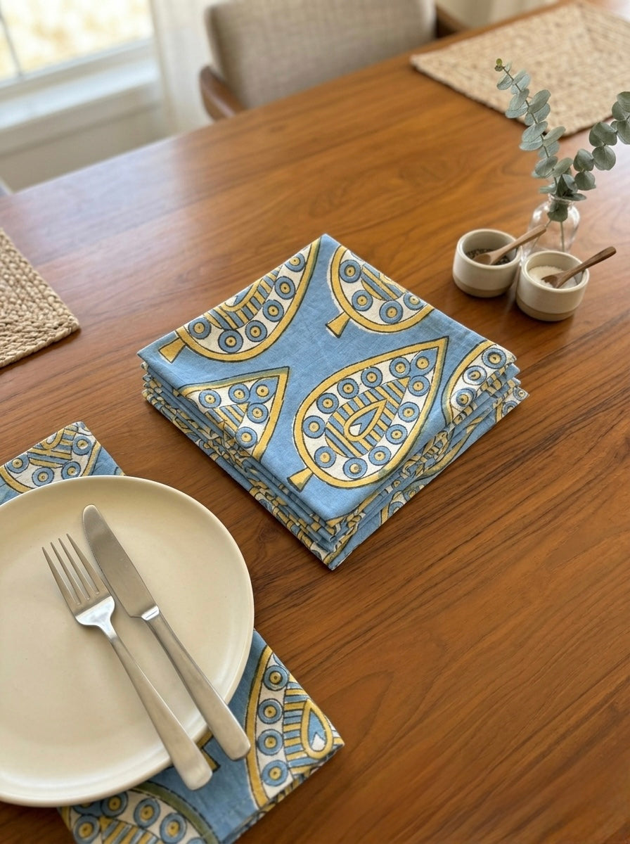 House of Mulmul - Blockprinted Cotton Table Napkins. Table setting with Set of blue napkins with gold leaf patterns, plates, and cutlery on a wooden table.