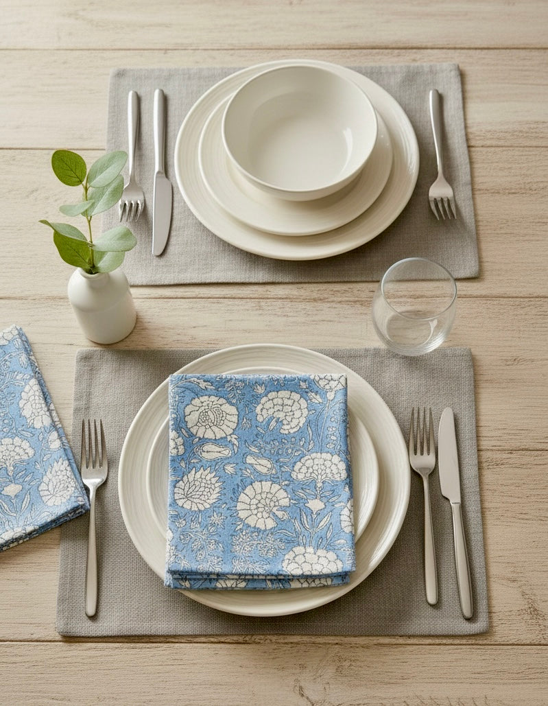 House of Mulmul - Blockprinted Cotton Napkins. Dining table setting with white plates, blue floral napkins, silverware, and a glass on a wooden surface.