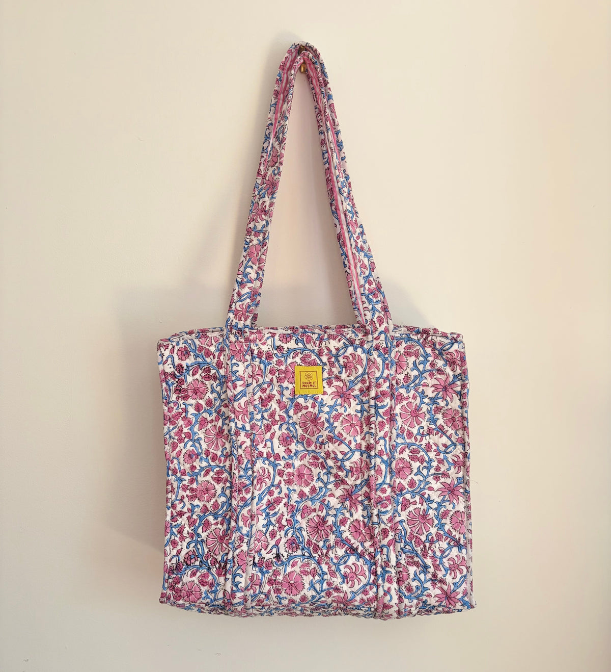 House of Mulmul - Blockprinted Cotton Tote Bag, Travel Bag. A quilted fabric tote bag with a pink, blue, and white floral pattern, featuring a small yellow square brand patch on the front, hanging against a plain beige wall.