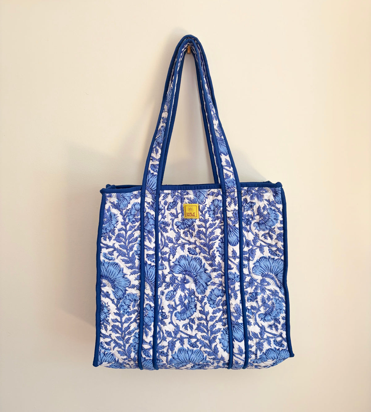 House of Mulmul - Blockprinted Cotton Tote Bag, Travel Bag. A blue and white floral print tote bag with blue handles and trim, hanging against a plain beige wall.