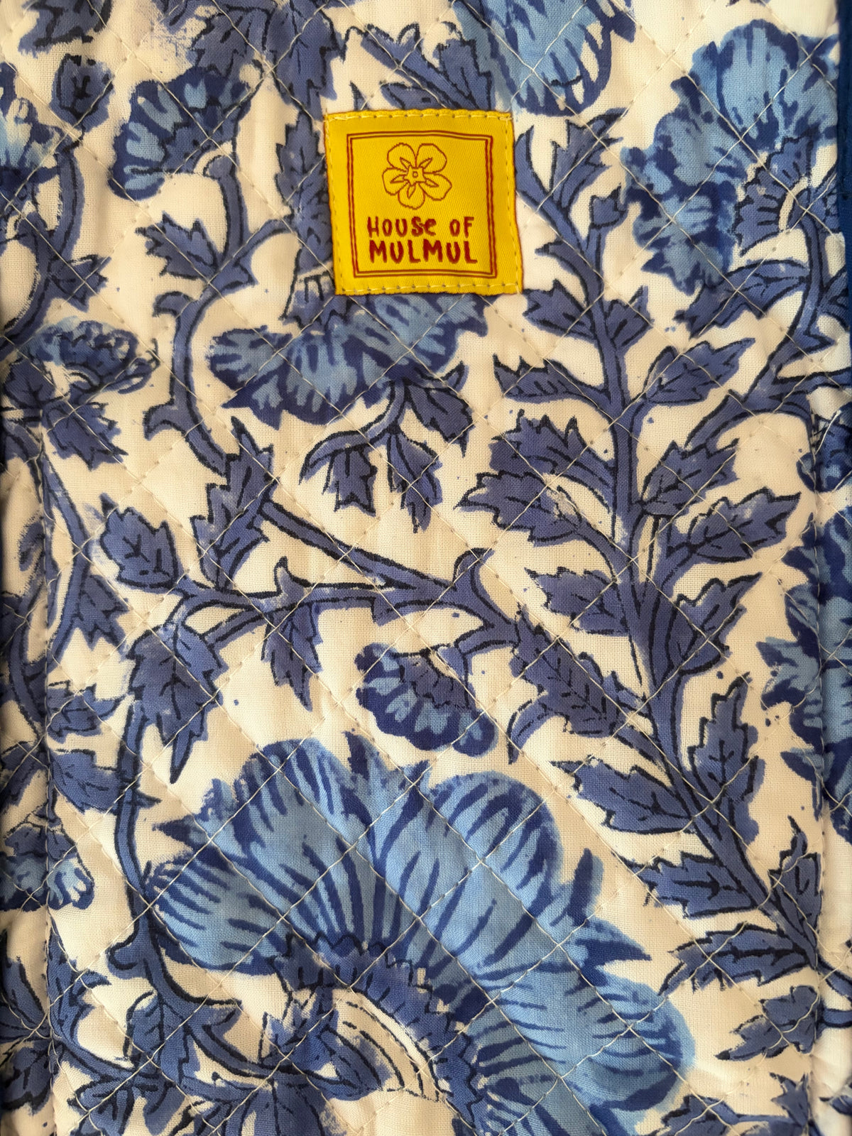 House of Mulmul - Blockprinted Cotton Tote Bag, Travel Bag. Close-up of a quilted textile featuring a blue floral vine pattern on a cream background, with a yellow square patch reading 'HOUSE OF MULMUL' near the top.