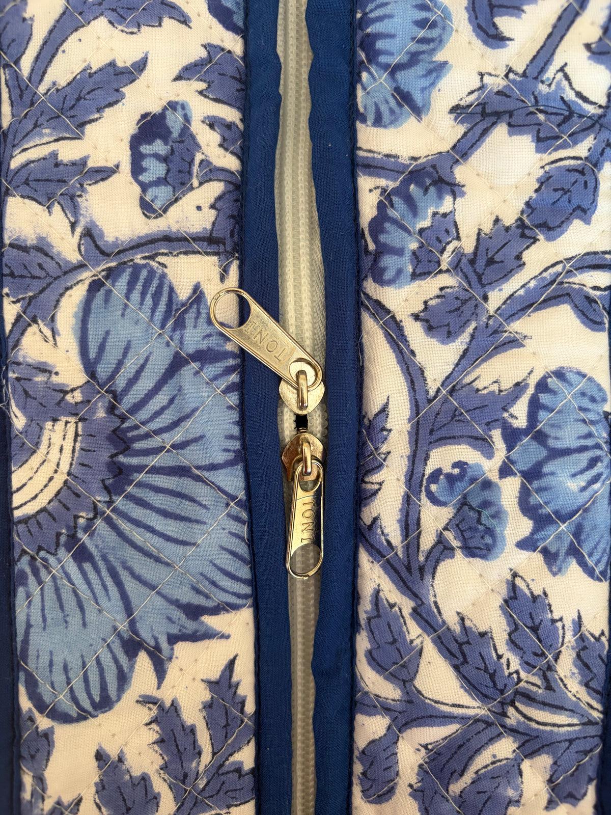 House of Mulmul - Blockprinted Cotton Tote Bag, Travel Bag. Close-up of a quilted fabric tote bag featuring a blue floral pattern on a cream background, with a central gold zipper.