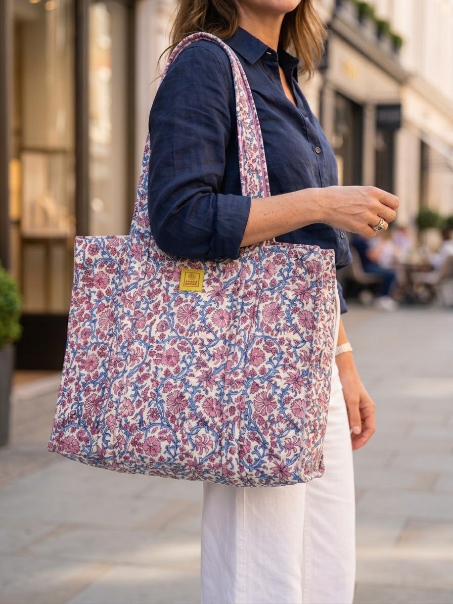 House of Mulmul - Blockprinted Cotton Tote Bag, Travel Bag. A woman wearing a navy shirt and white trousers carries a large floral tote bag with pink, purple, and blue flowers on a light background, standing on a city street.