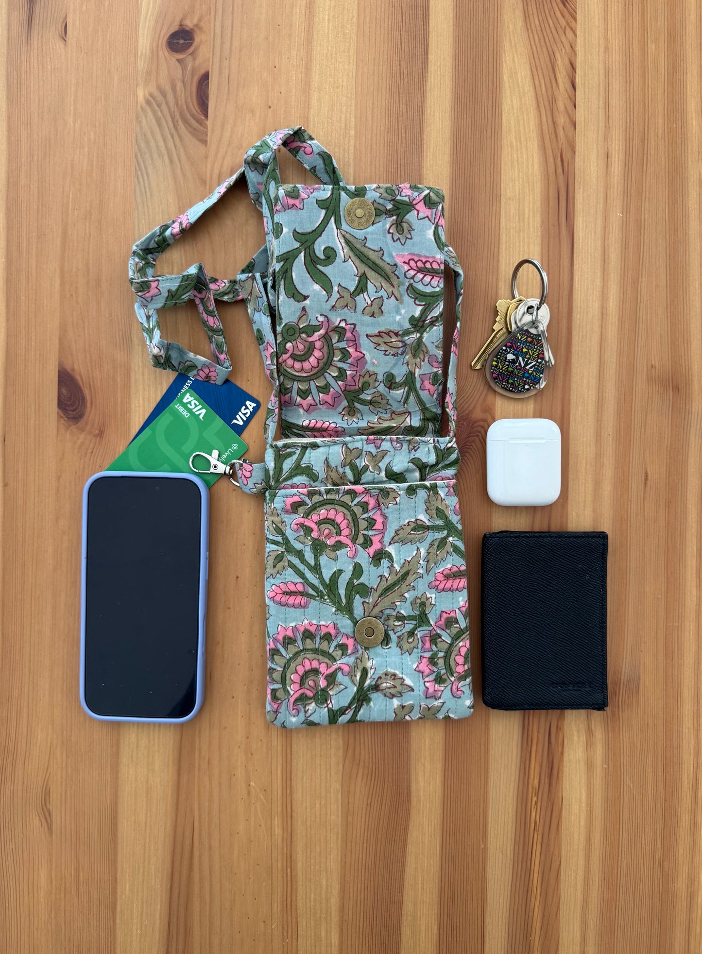 House of Mulmul - Blockprinted Crossbody Mobile Sling Bag. Floral crossbody bag with phone, cards, wallet, keys, and airpods on a wooden surface