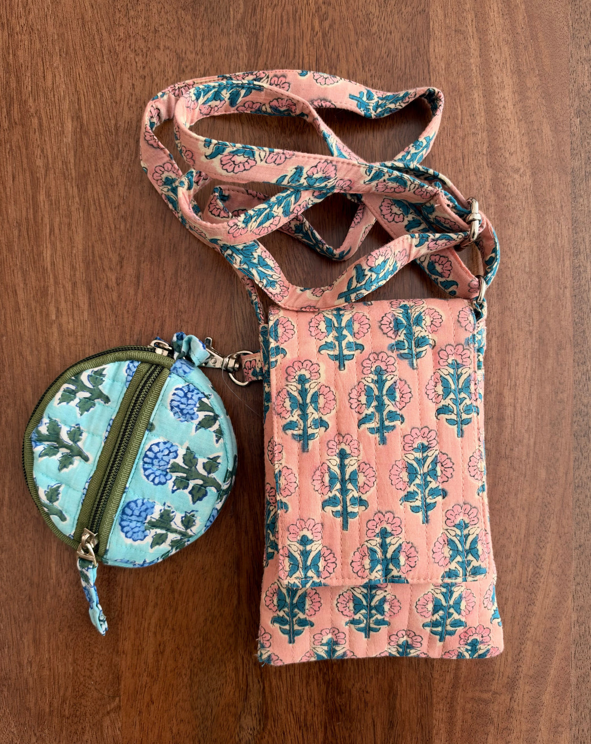 House of Mulmul - Blockprinted Mobile Sling Bag. A peach-colored fabric crossbody phone bag with a teal and pink block print pattern, shown with a matching round coin pouch on a brown wooden background.