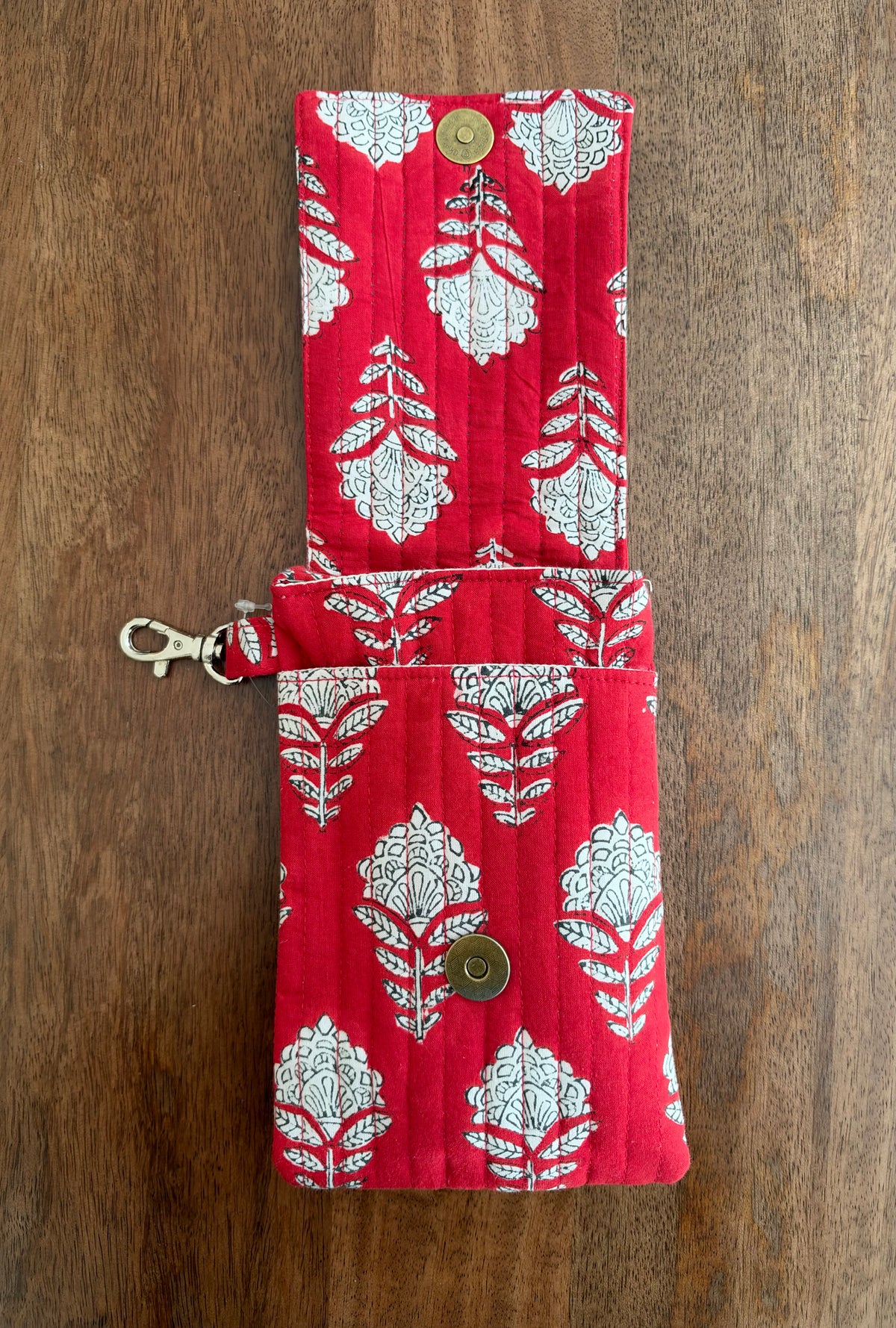 House of Mulmul - Blockprinted Mobile Sling Bag. A red quilted fabric pouch with a white floral block print pattern, featuring a snap button closure and a metal lobster clasp, laid flat on a wooden surface.