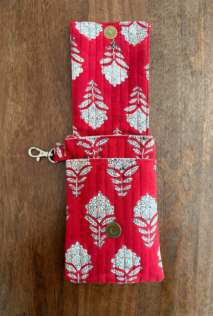 House of Mulmul - Blockprinted Mobile Sling Bag. A red quilted fabric pouch with a white floral block print pattern, featuring a snap button closure and a metal lobster clasp, laid flat on a wooden surface.