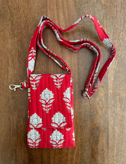 House of Mulmul - Blockprinted Mobile Sling Bag. A red quilted fabric crossbody bag with a white floral block print pattern and a matching adjustable shoulder strap, laid flat on a brown wooden surface.