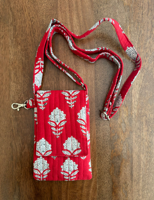 House of Mulmul - Blockprinted Mobile Sling Bag. A red quilted fabric crossbody bag with a white floral block print pattern and a matching adjustable shoulder strap, laid flat on a brown wooden surface.