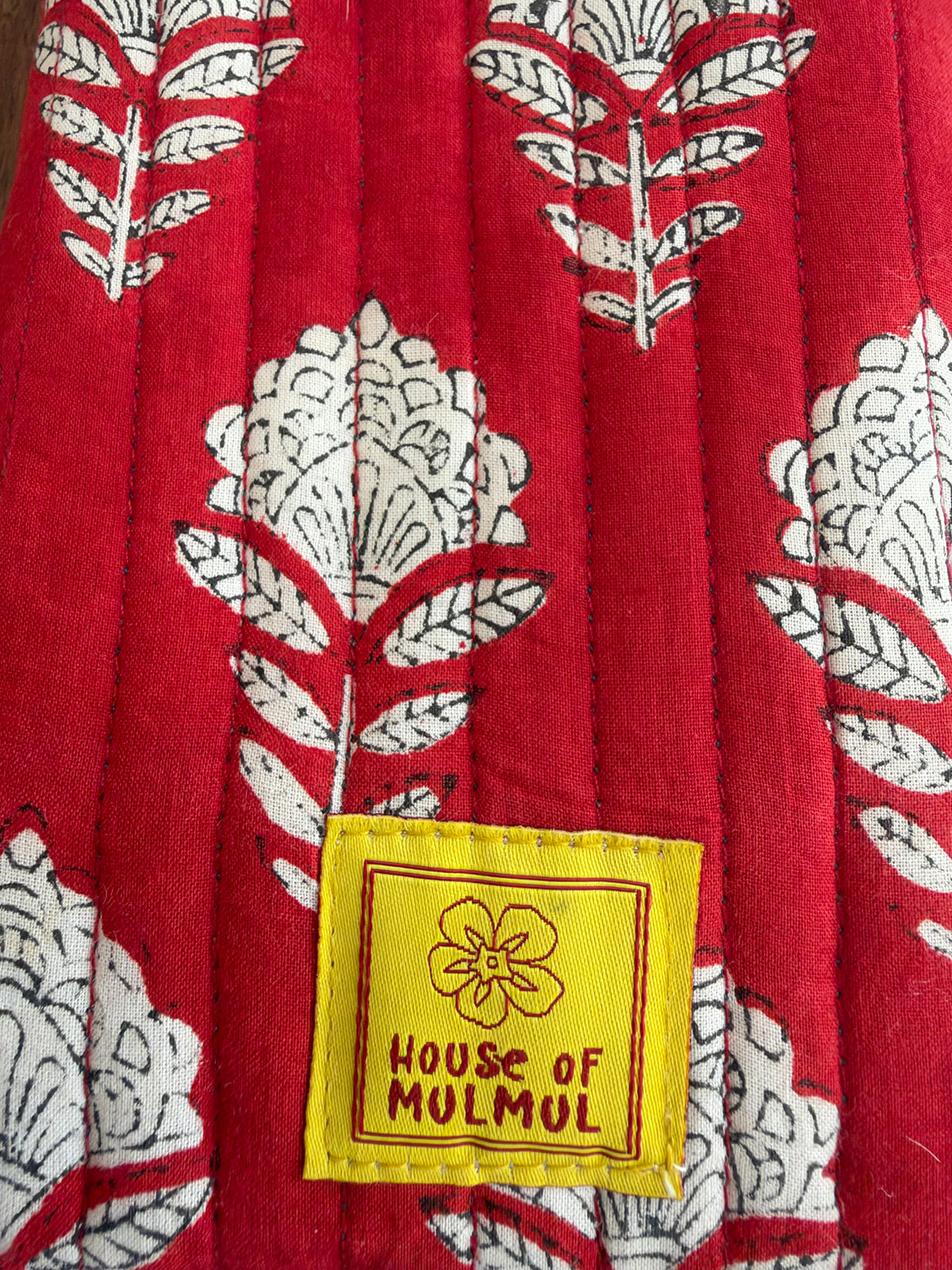 House of Mulmul - Blockprinted Mobile Sling Bag. Close-up of a red quilted textile with a white floral and leaf pattern and a yellow sewn label reading 'HOUSE OF MULMUL'.