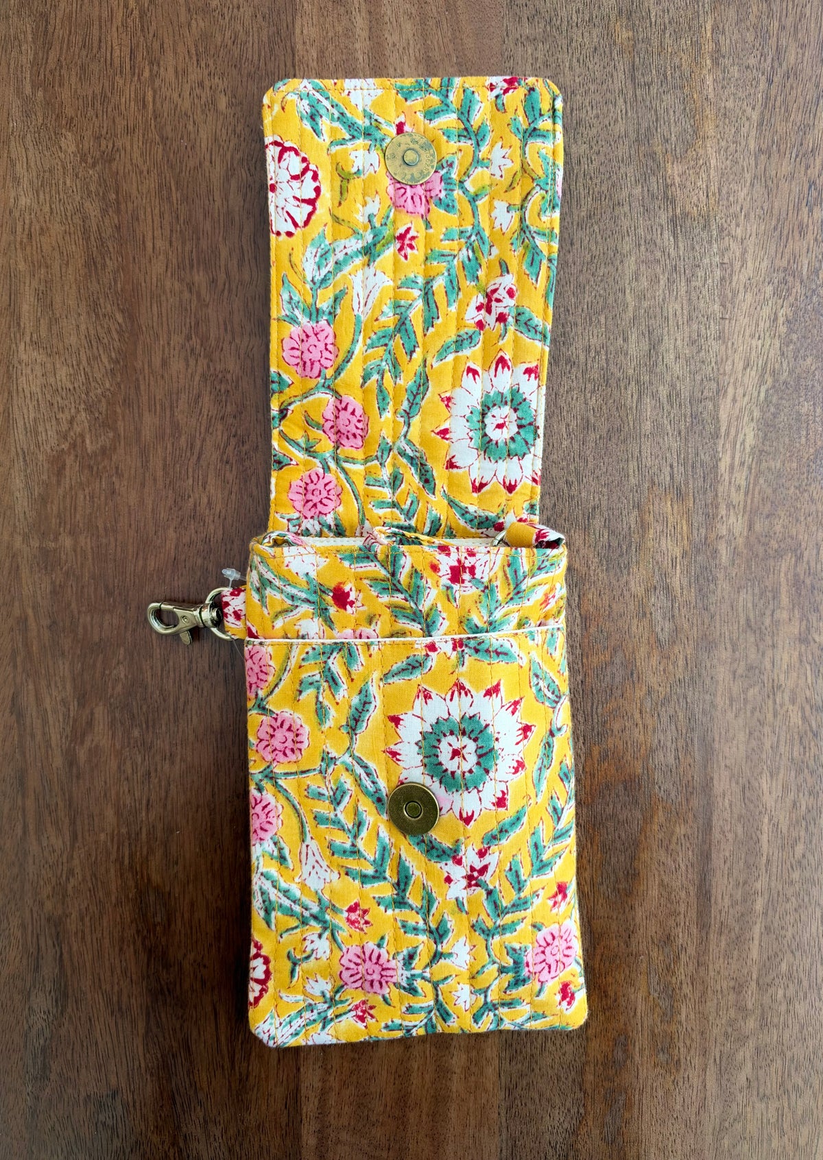 House of Mulmul - Blockprinted Mobile Sling Bag. A yellow fabric phone pouch with a colorful floral and leaf pattern, featuring a snap button closure and a metal ring detail, shown on a dark wood background.