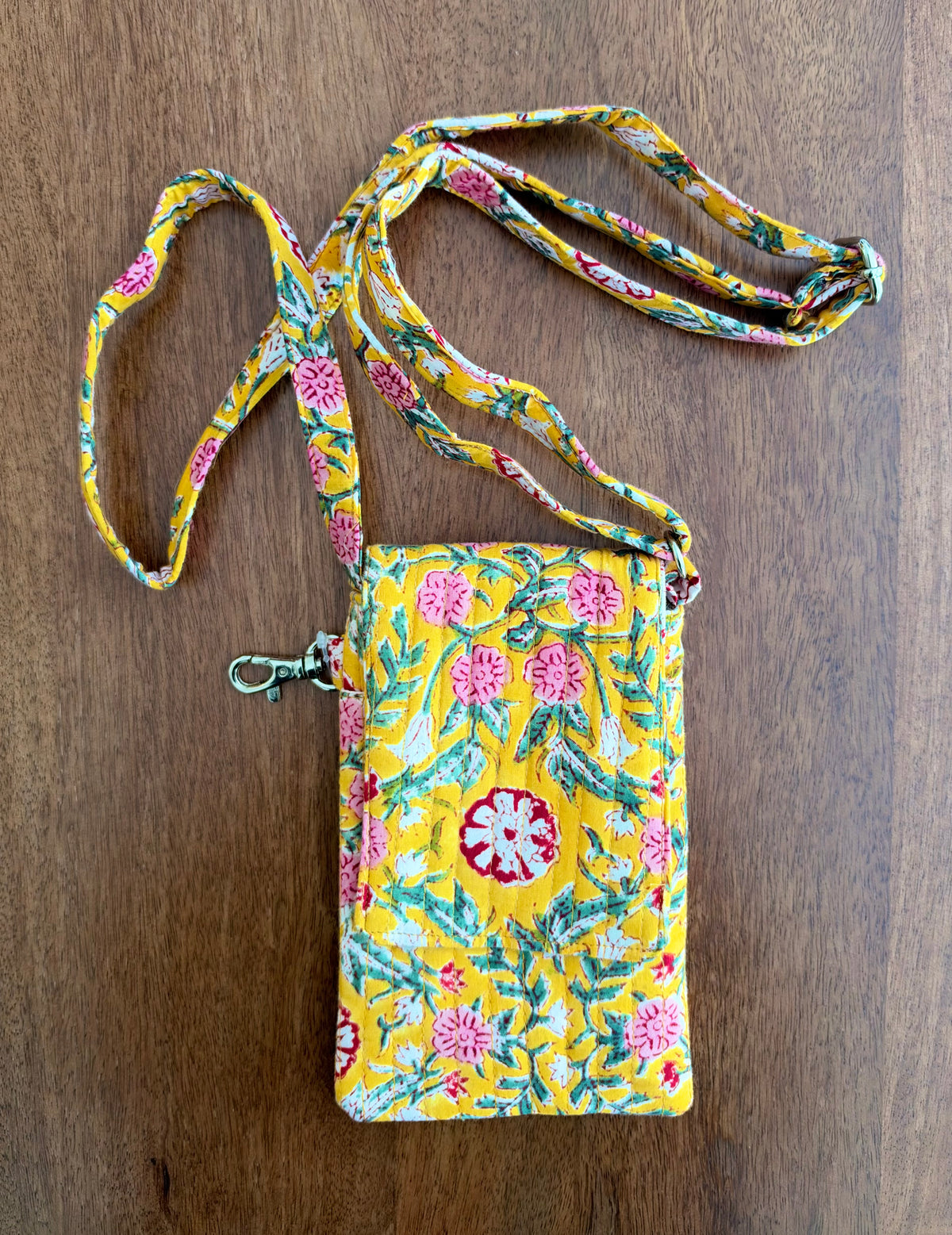 House of Mulmul - Blockprinted Mobile Sling Bag. A yellow fabric crossbody phone pouch with a pink and green floral pattern, featuring a matching adjustable strap and a silver metal clasp, laid flat on a wooden surface.