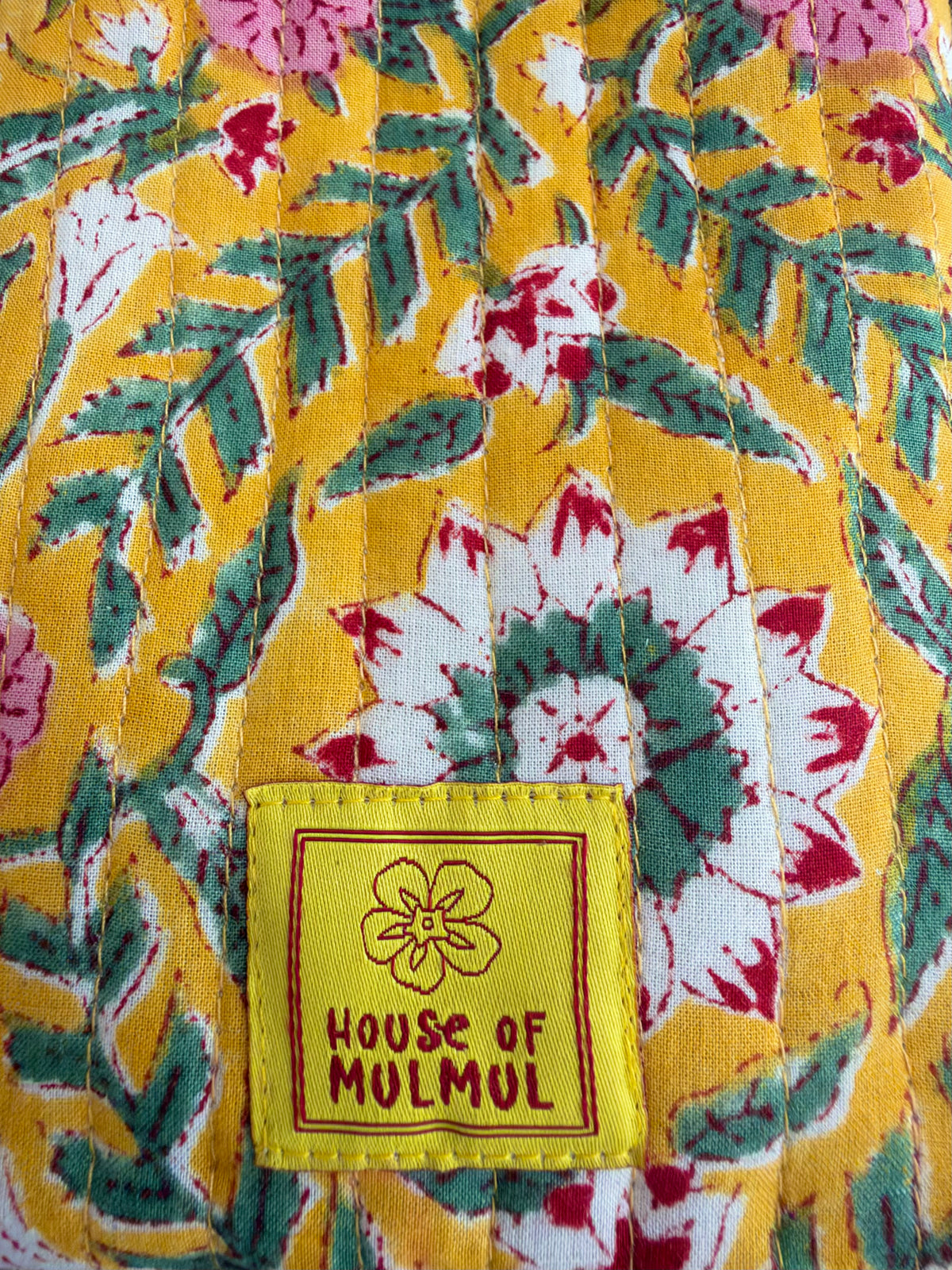 House of Mulmul - Blockprinted Mobile Sling Bag. Close-up of a quilted fabric with a yellow base and a multicolor floral pattern of green leaves, white and pink flowers, and a yellow sewn-on brand patch reading 'HOUSE OF MULMUL'.