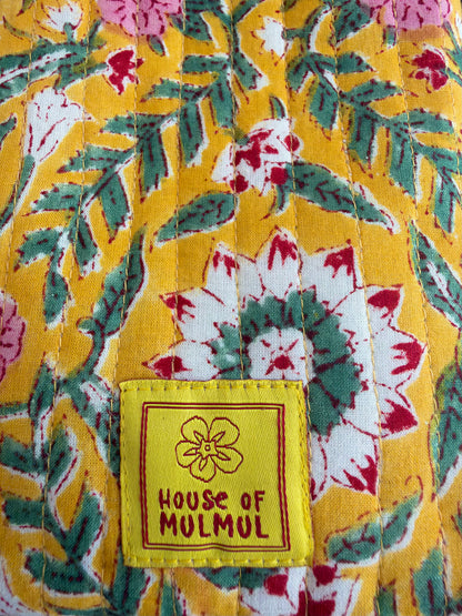 House of Mulmul - Blockprinted Mobile Sling Bag. Close-up of a quilted fabric with a yellow base and a multicolor floral pattern of green leaves, white and pink flowers, and a yellow sewn-on brand patch reading 'HOUSE OF MULMUL'.