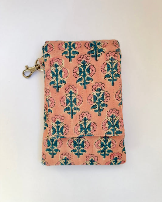 House of Mulmul - Blockprinted Mobile Sling Bag. A small fabric mobile pouch or wallet with a peach-pink background and a repeating block-print style floral pattern in teal and coral pink, featuring a brass clasp on the left side, photographed against a plain off-white background.