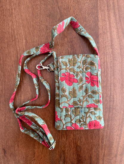 House of Mulmul - Blockprinted Mobile Sling Bag. A fabric crossbody phone pouch with a floral print, featuring a light blue-green background with pink flowers and brown leaves, complete with a matching adjustable strap and metal clasp, laid flat on a wooden surface.