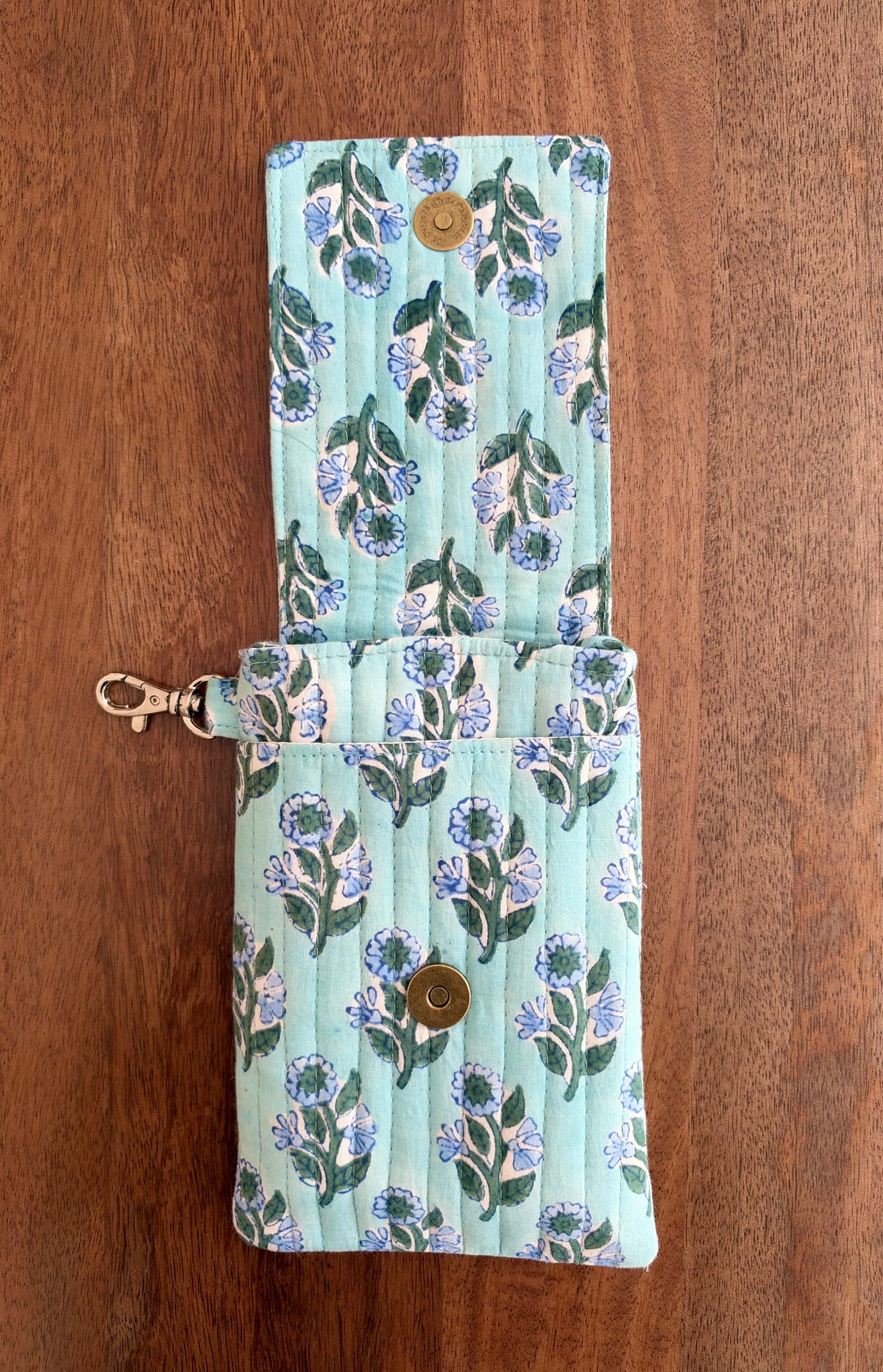 House of Mulmul - Blockprinted Mobile Sling Bag. A quilted fabric wallet in a light mint green color with a blue and green floral pattern, featuring a snap button closure and a brass clasp, displayed on a wooden surface.