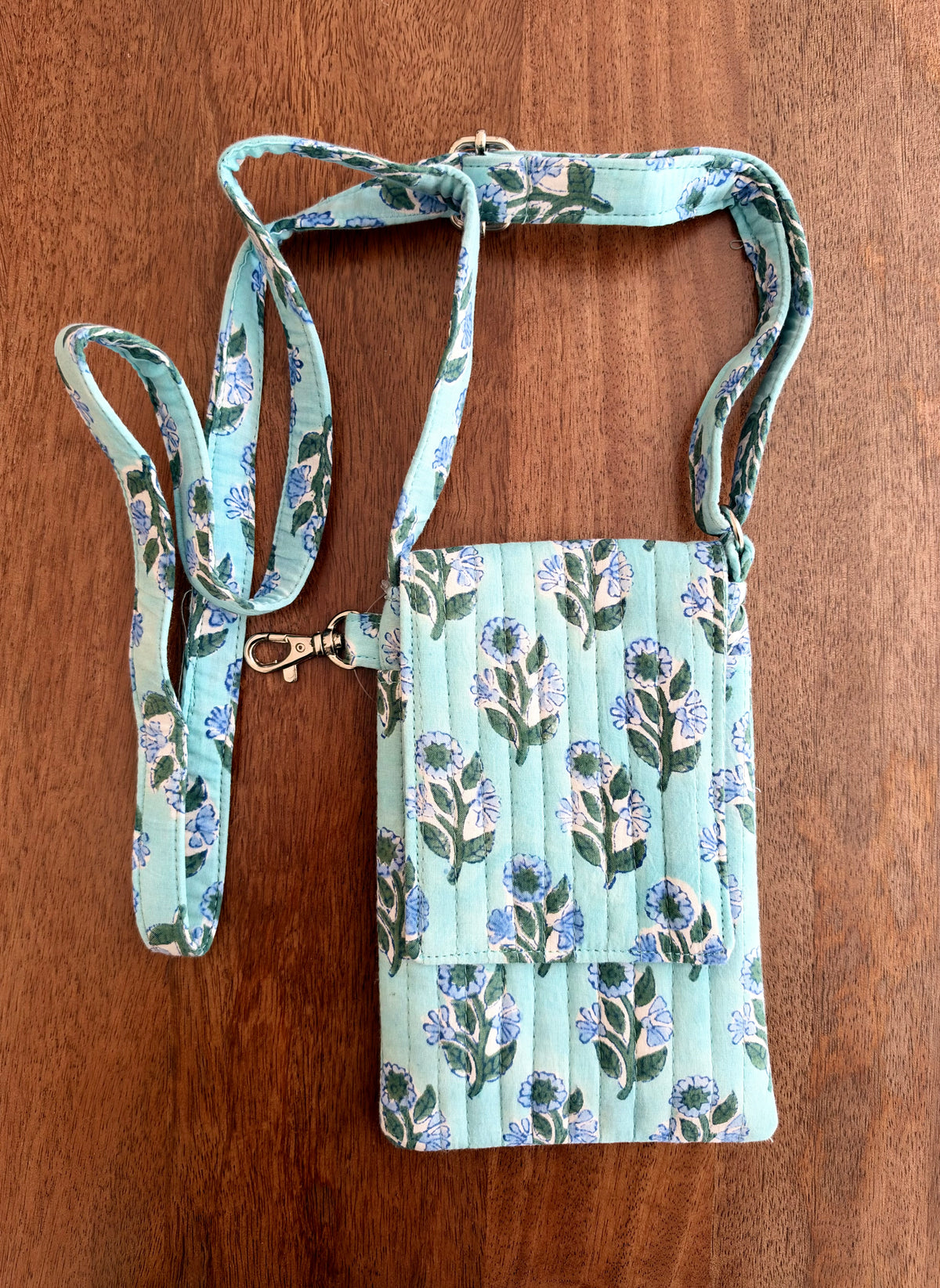House of Mulmul - Blockprinted Mobile Sling Bag. A small quilted fabric crossbody phone bag with a floral print in mint green, blue, and white, shown on a brown wooden background with its matching strap laid out beside it.