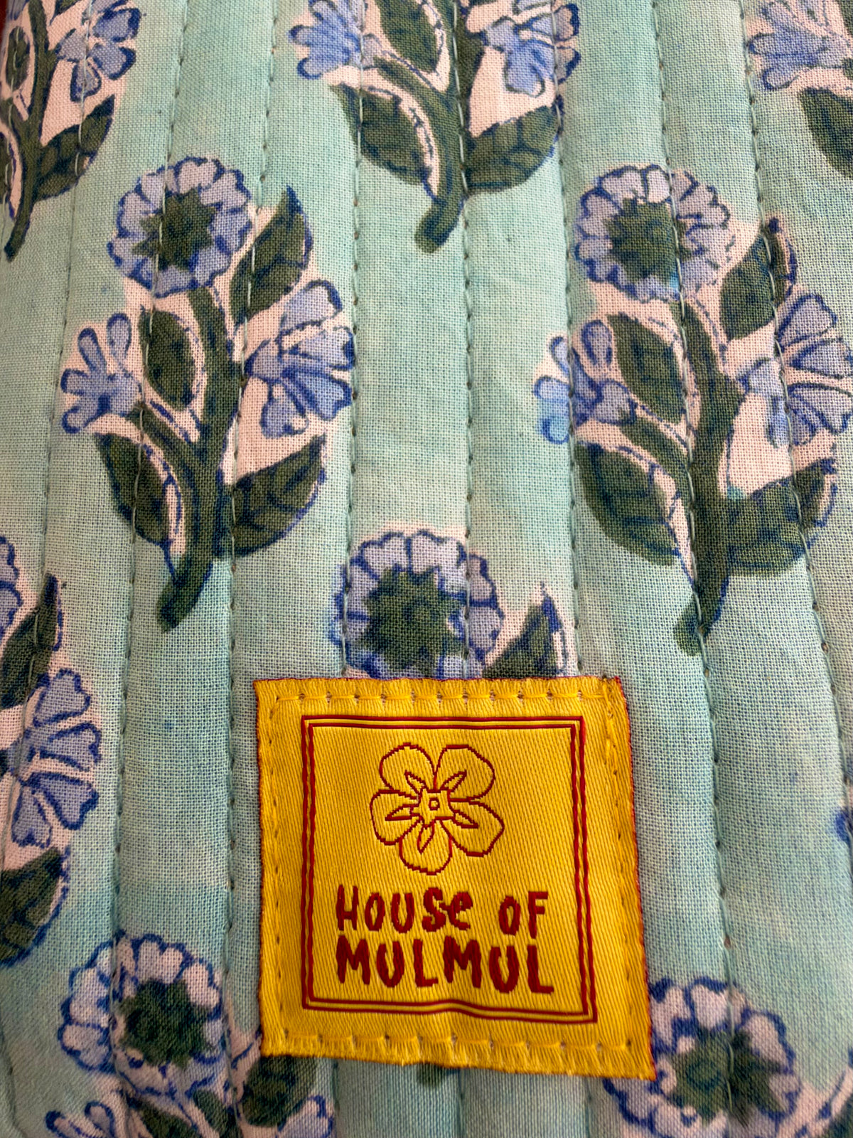 House of Mulmul - Blockprinted Mobile Sling Bag. Close-up of a quilted fabric with a light blue-green background and a floral block print in blue, green, and off-white. A yellow sewn-on brand label reading 'HOUSE OF MULMUL' is visible near the bottom center.