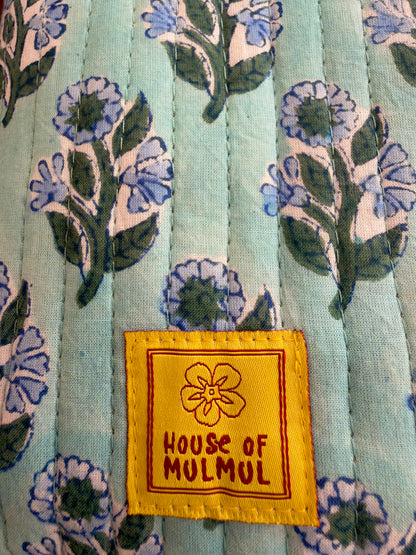 House of Mulmul - Blockprinted Mobile Sling Bag. Close-up of a quilted fabric with a light blue-green background and a floral block print in blue, green, and off-white. A yellow sewn-on brand label reading 'HOUSE OF MULMUL' is visible near the bottom center.
