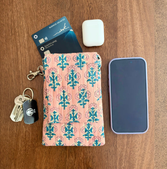 House of Mulmul - Blockprinted Mobile Sling Bag. A pink patterned mobile sling bag with a blue and white tree motif, shown flat-laid on a wooden table alongside a smartphone, keys, a credit card, and a white earbud case.