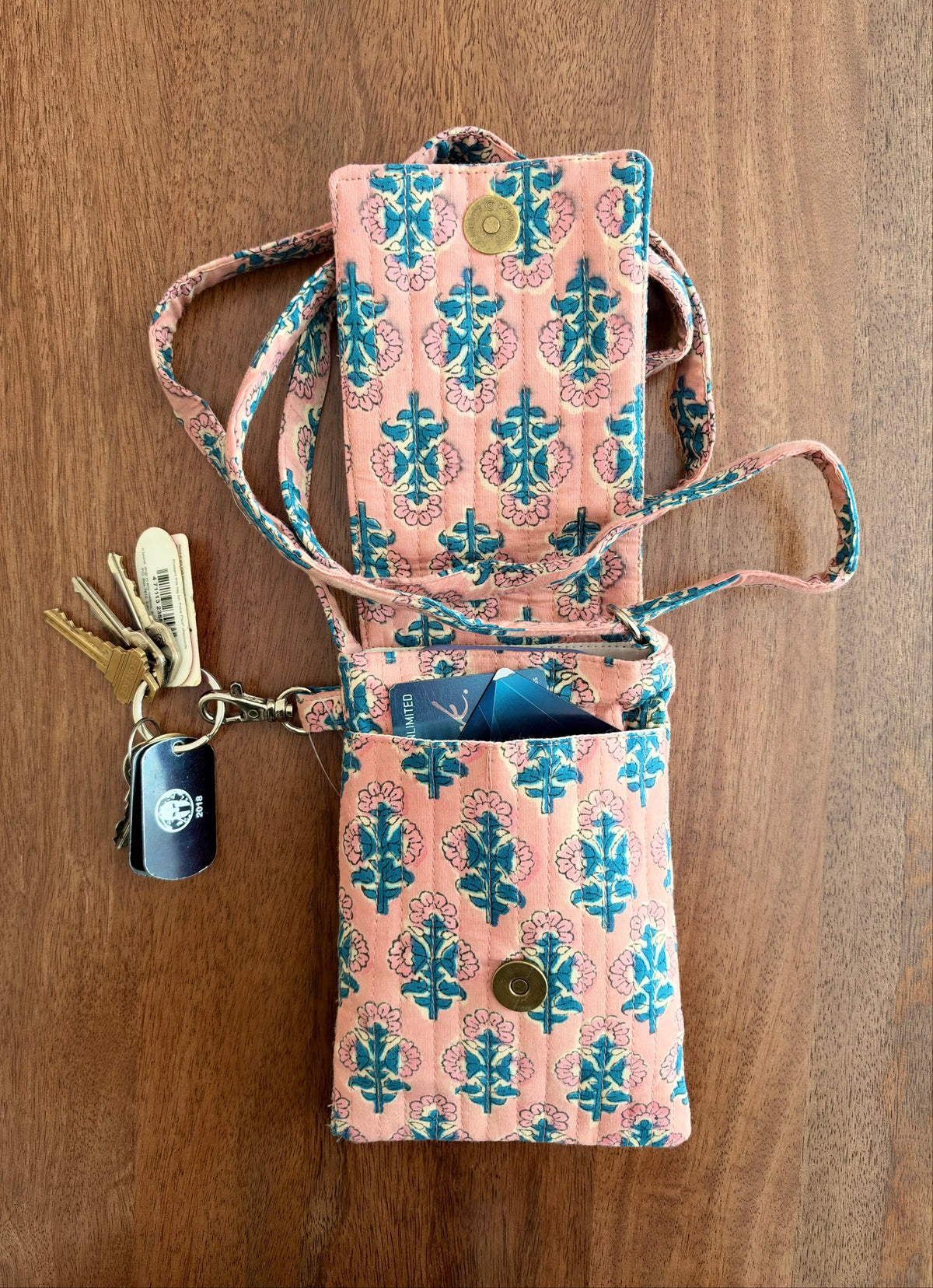 House of Mulmul - Blockprinted Mobile Sling Bag. A pink and teal patterned mobile sling bag holder wallet with a strap, shown open on a wooden surface with keys and cards inside.