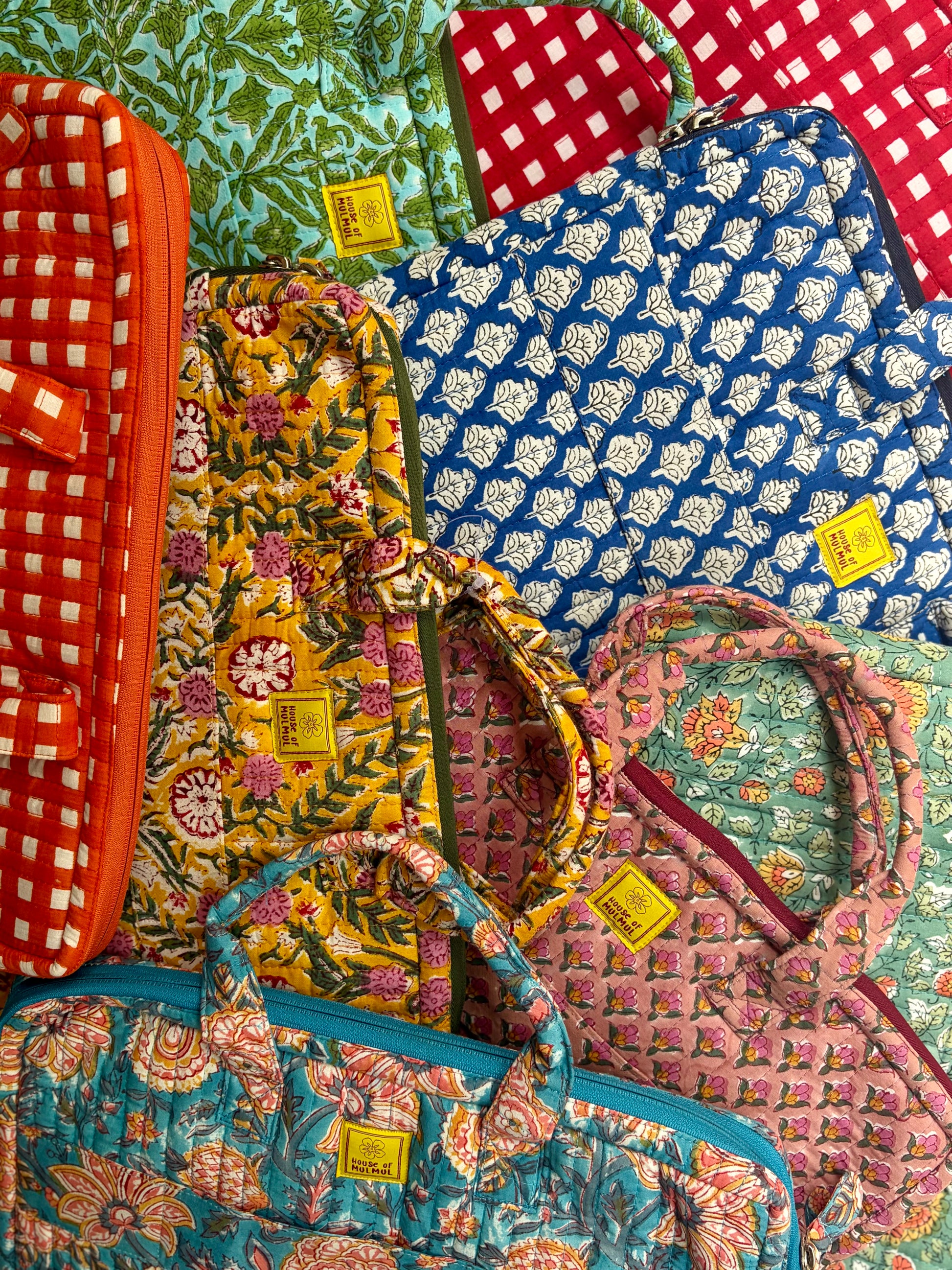 House of Mulmul - Blockprinted Cotton Quilted Laptop Bag. A collection of colorful quilted fabric bags and cases with various floral, geometric, and checkered patterns, arranged in a flat-lay composition. Each bag features a small yellow brand tag.